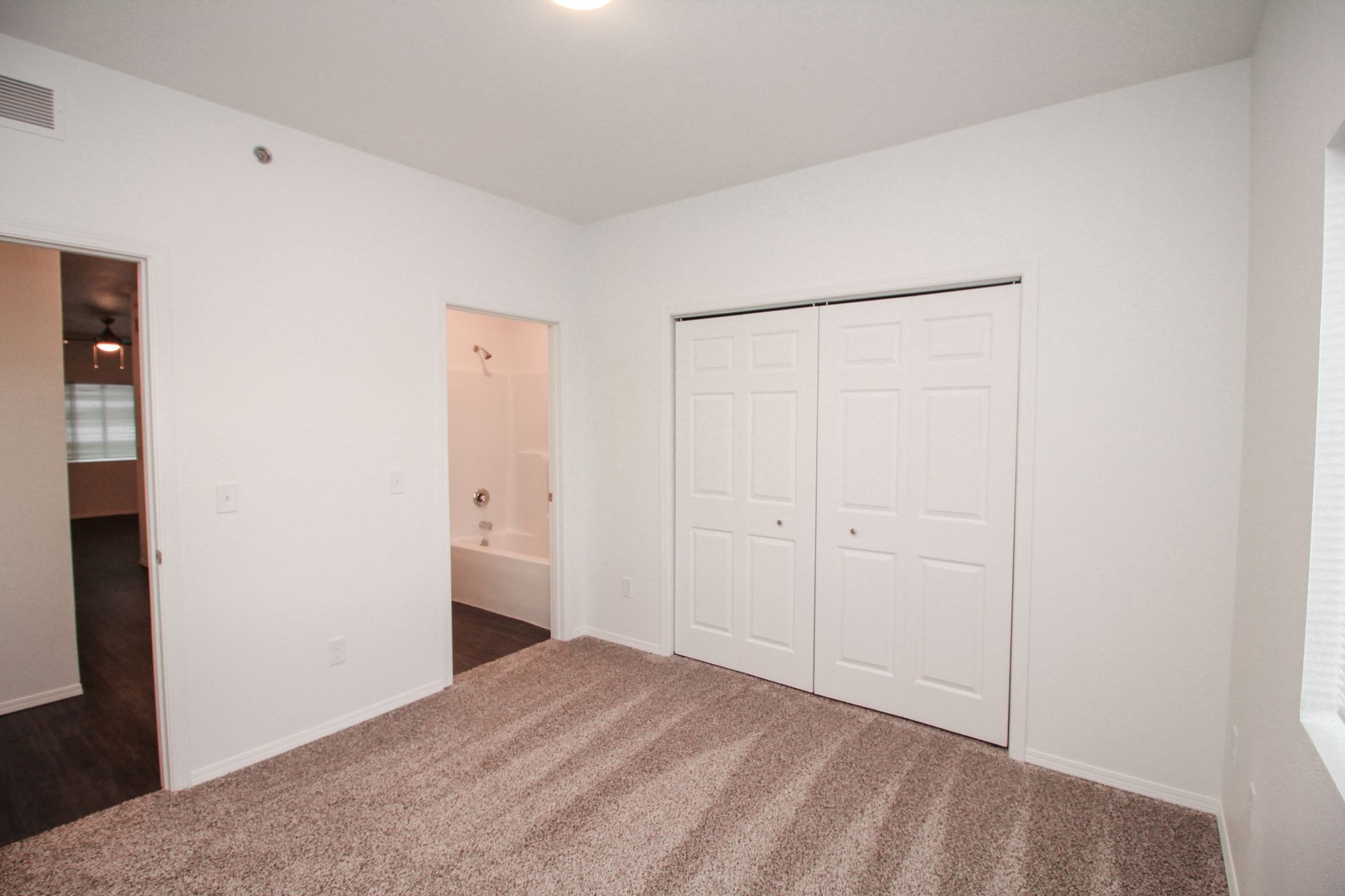 Empty white bedroom with beige carpet, closet doors, and an open doorway to a bathroom.