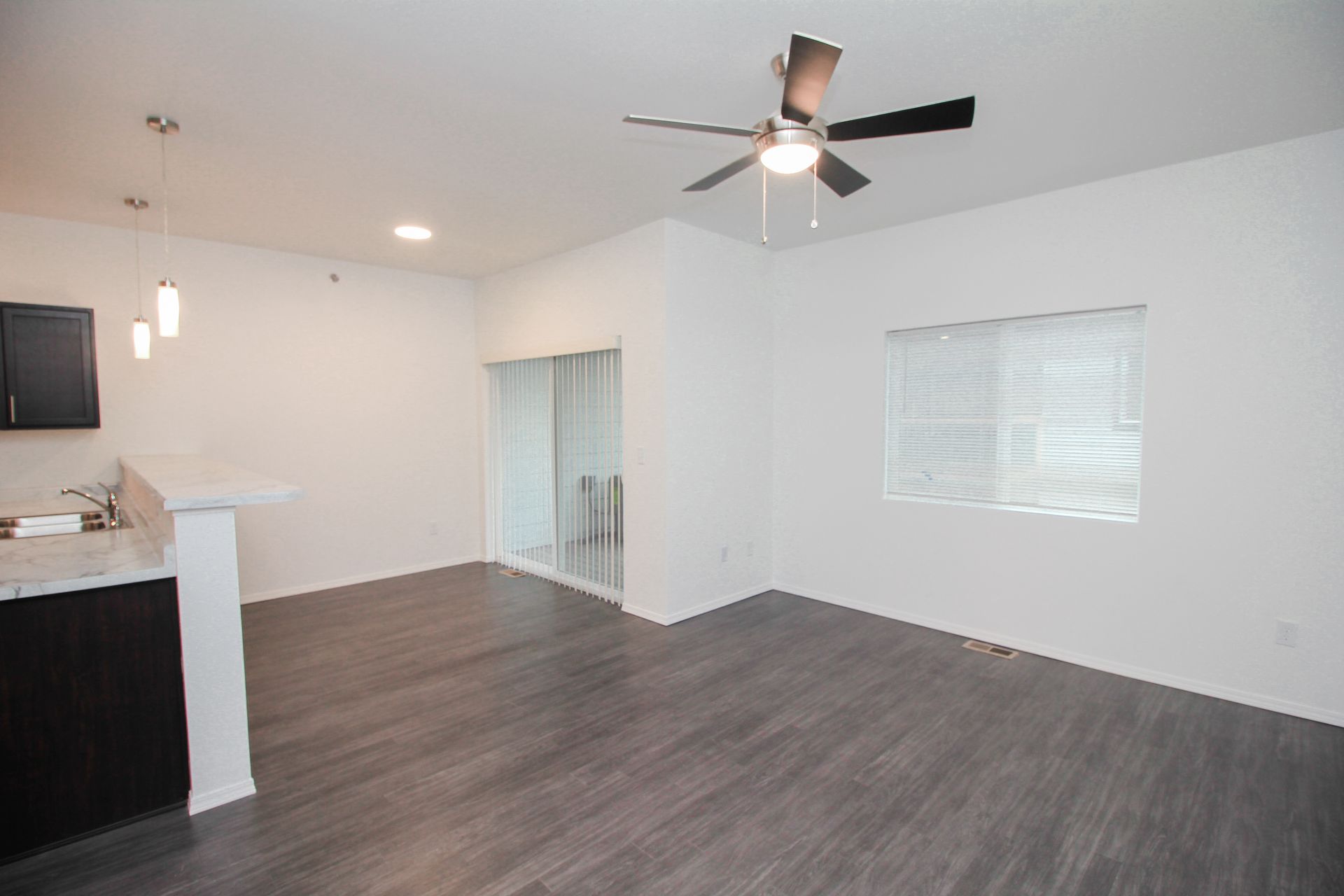 Empty modern apartment living room with dark wood floors, ceiling fan, and open kitchen with breakfast bar.