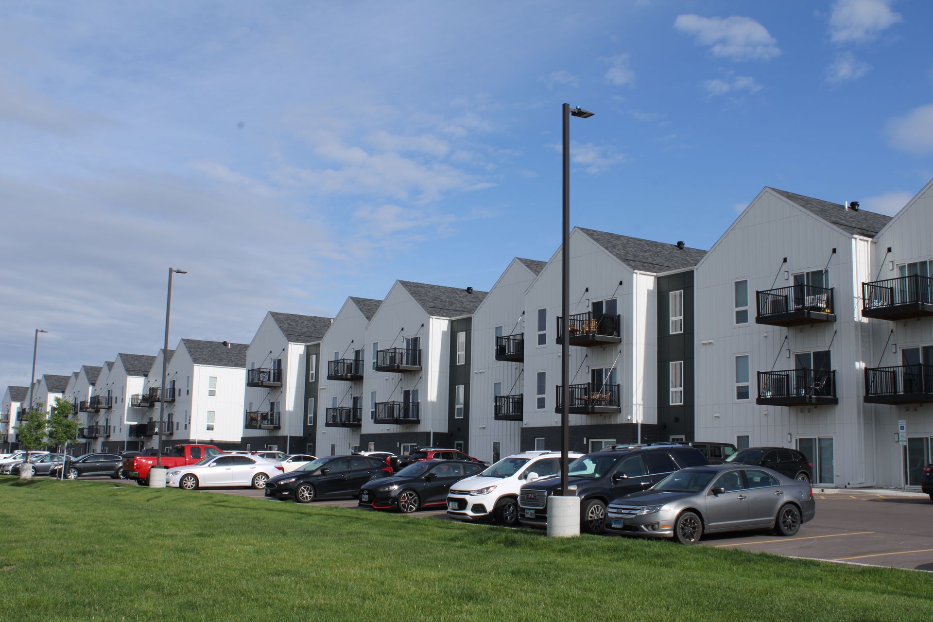 Modern white apartment buildings with parked cars under a blue sky along a grassy roadside