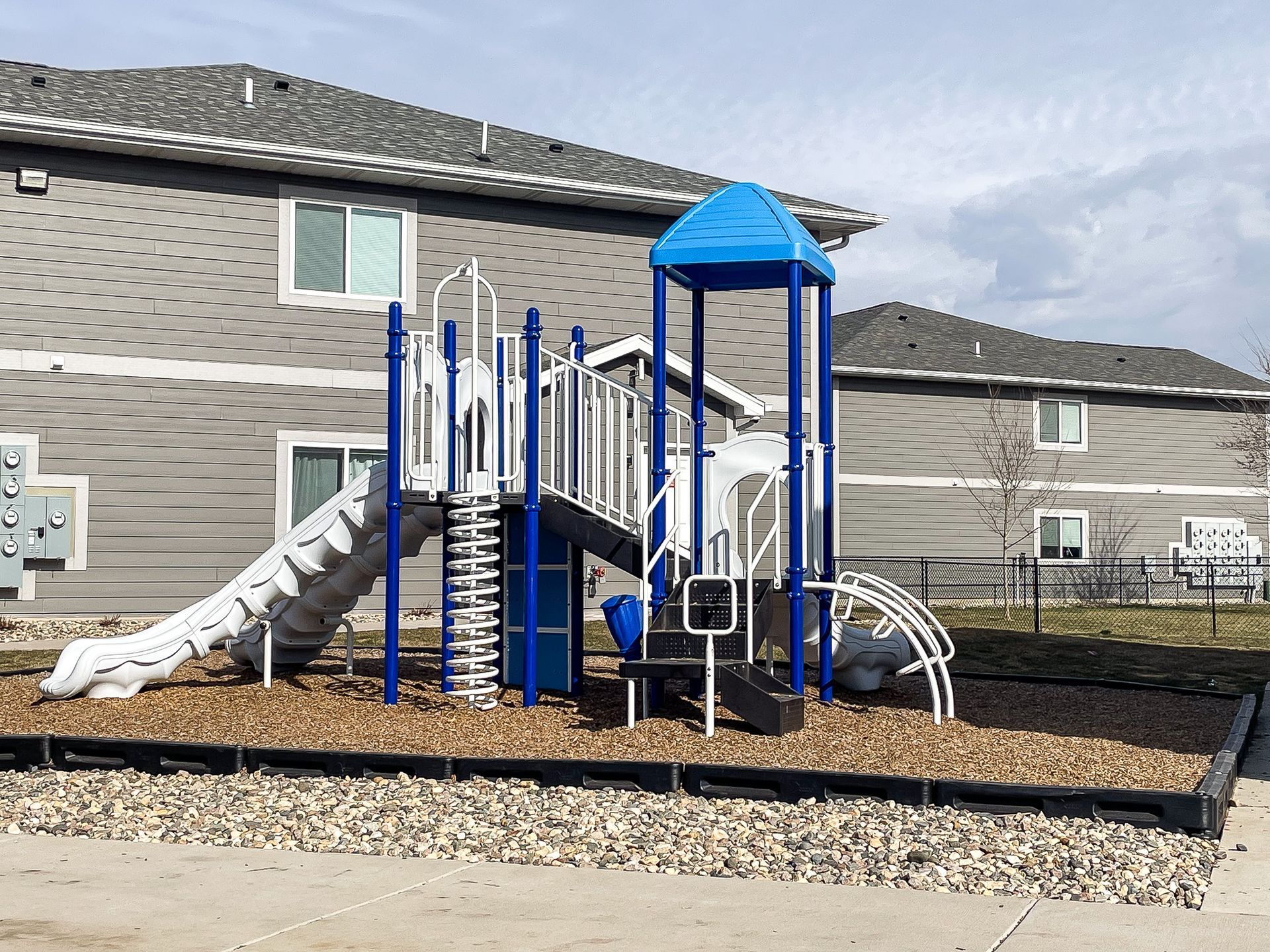 Playground play structure with blue roof and slides on a mulch surface outside a residential building