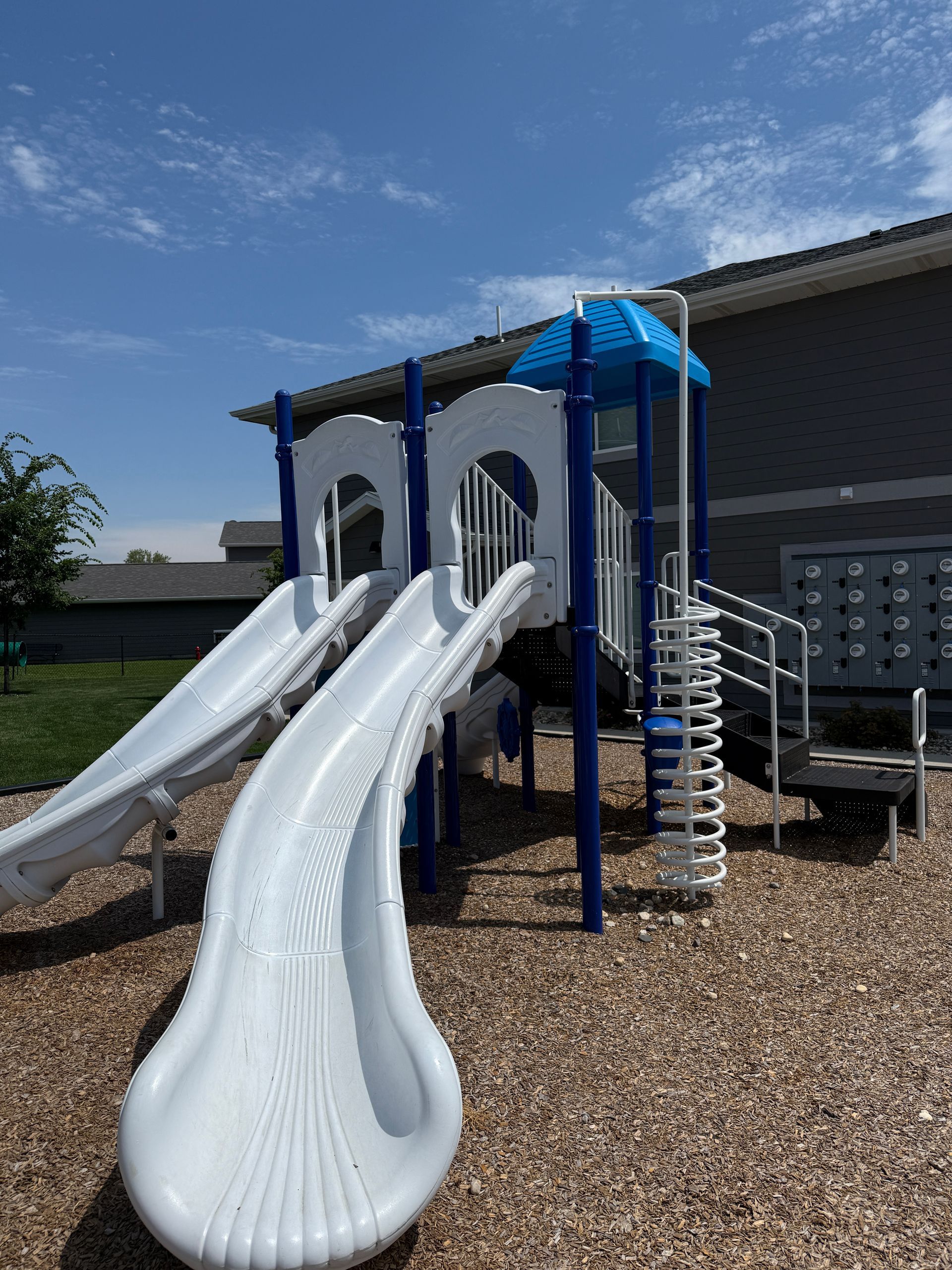 Playground slide structure with two white slides and blue roof on a gravel surface under a sunny sky
