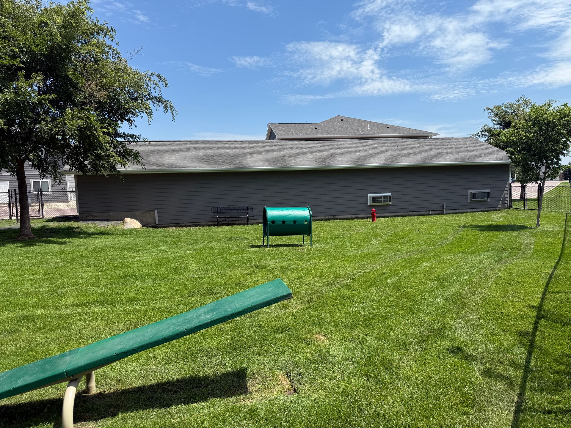 Green backyard with a wooden beam balance obstacle and a small green tunnel near a gray building.