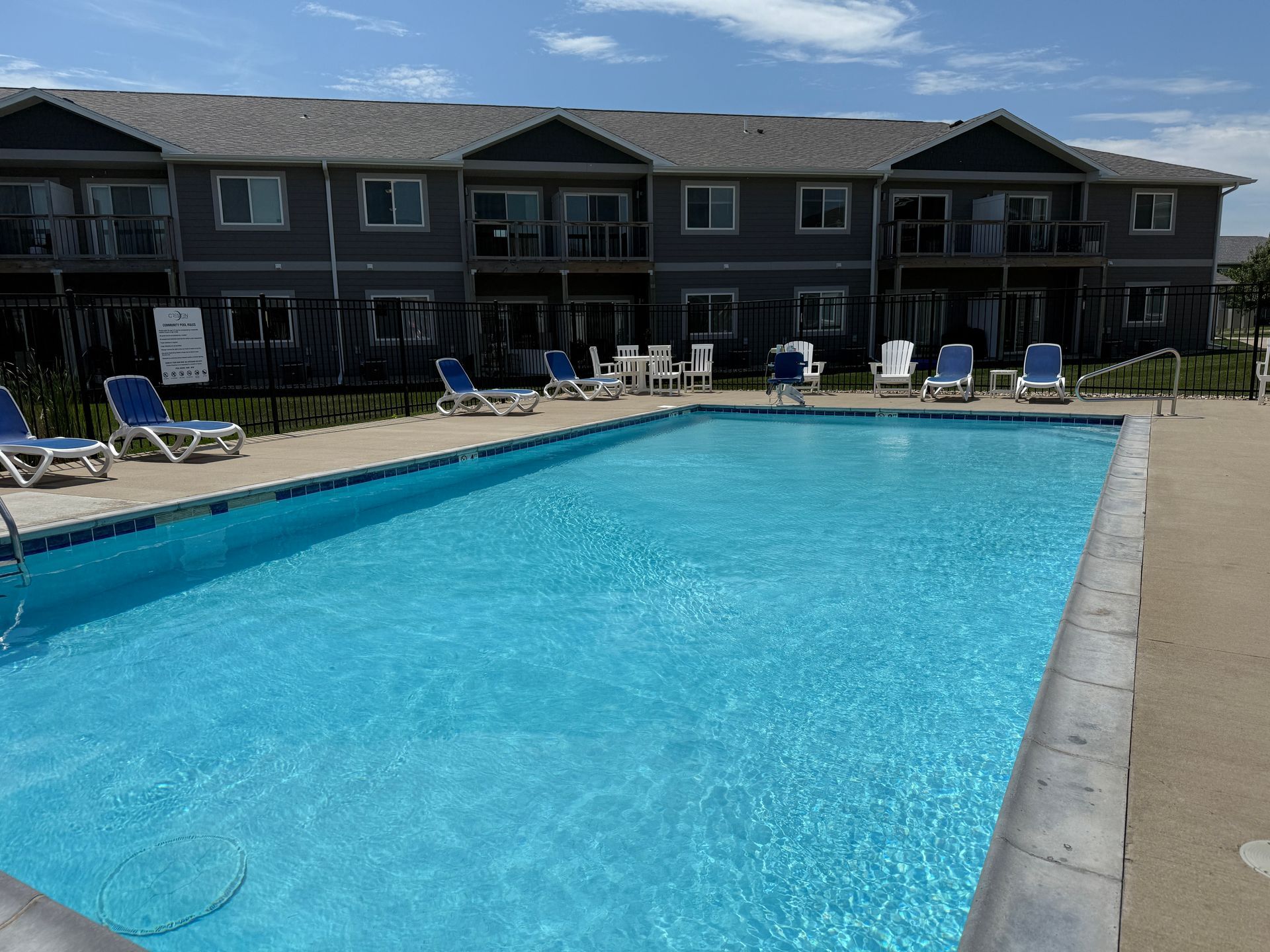 Outdoor swimming pool with blue water and lounge chairs beside a two-story apartment building.