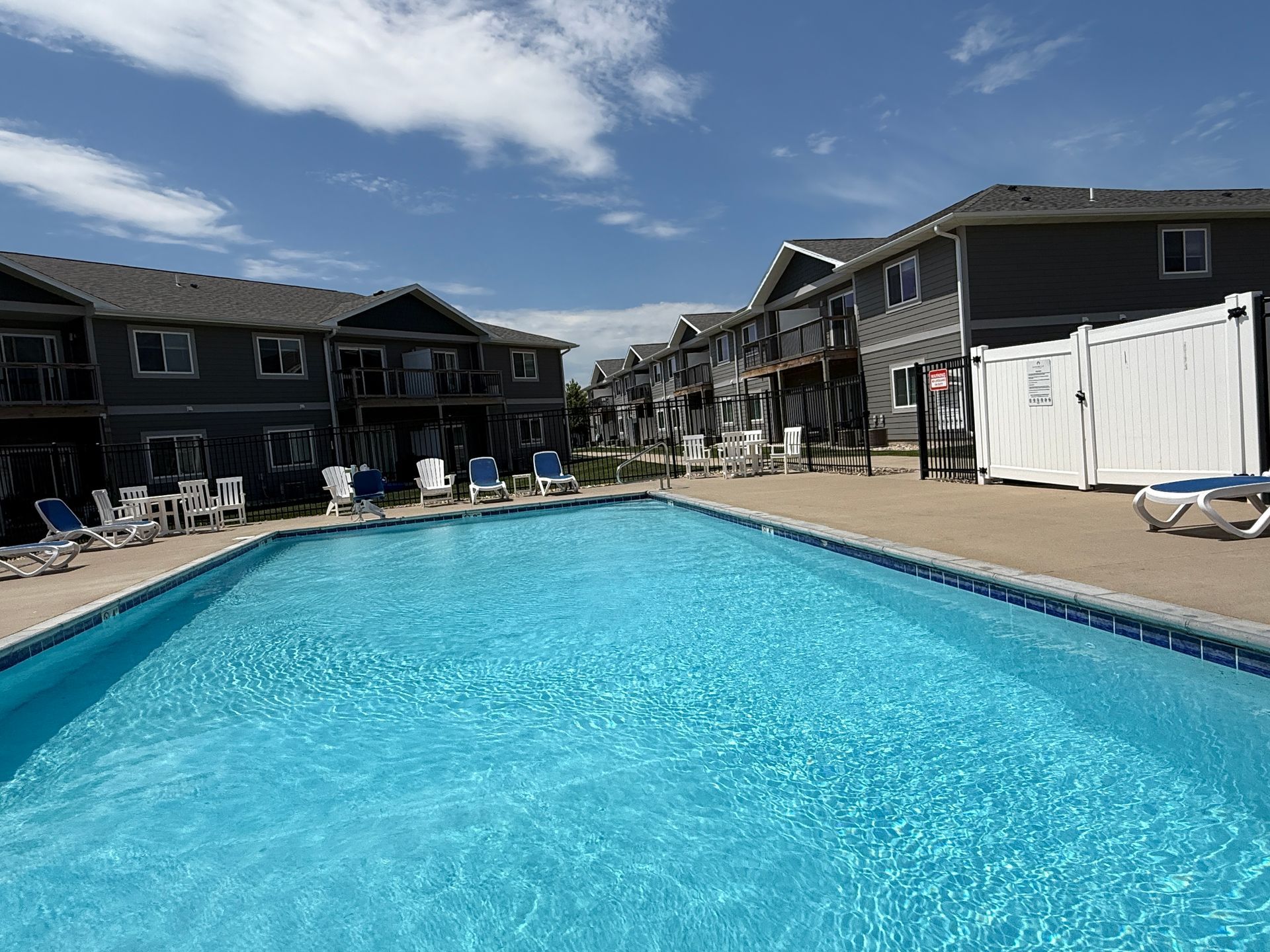 Outdoor swimming pool between resort buildings under a blue sky, with lounge chairs and white fence.