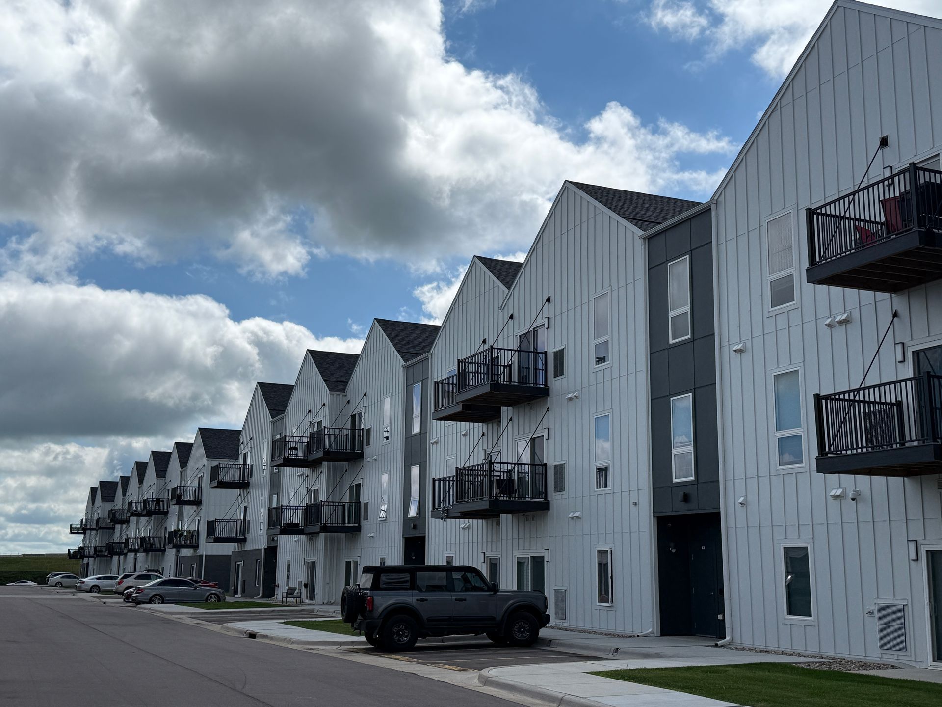 Row of white apartment buildings with black balconies under a cloudy sky, parked SUV in front