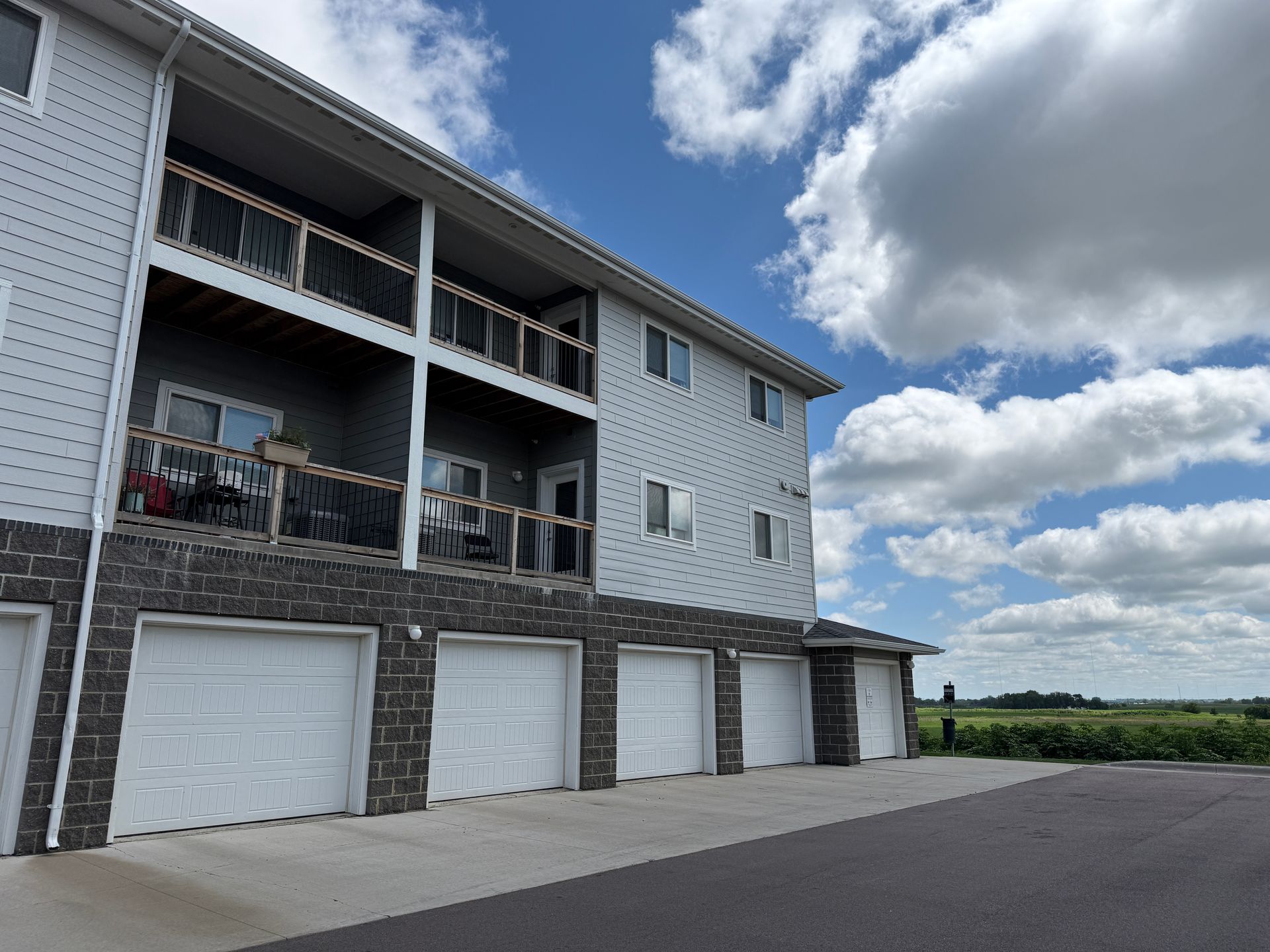 Modern apartment building with balconies and garage doors beside a large paved lot under a cloudy sky