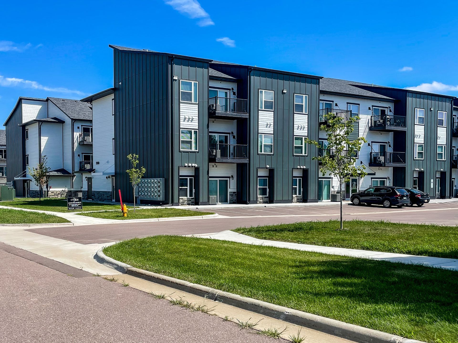 Modern gray apartment buildings beside a curved road and grassy lawn under a blue sky