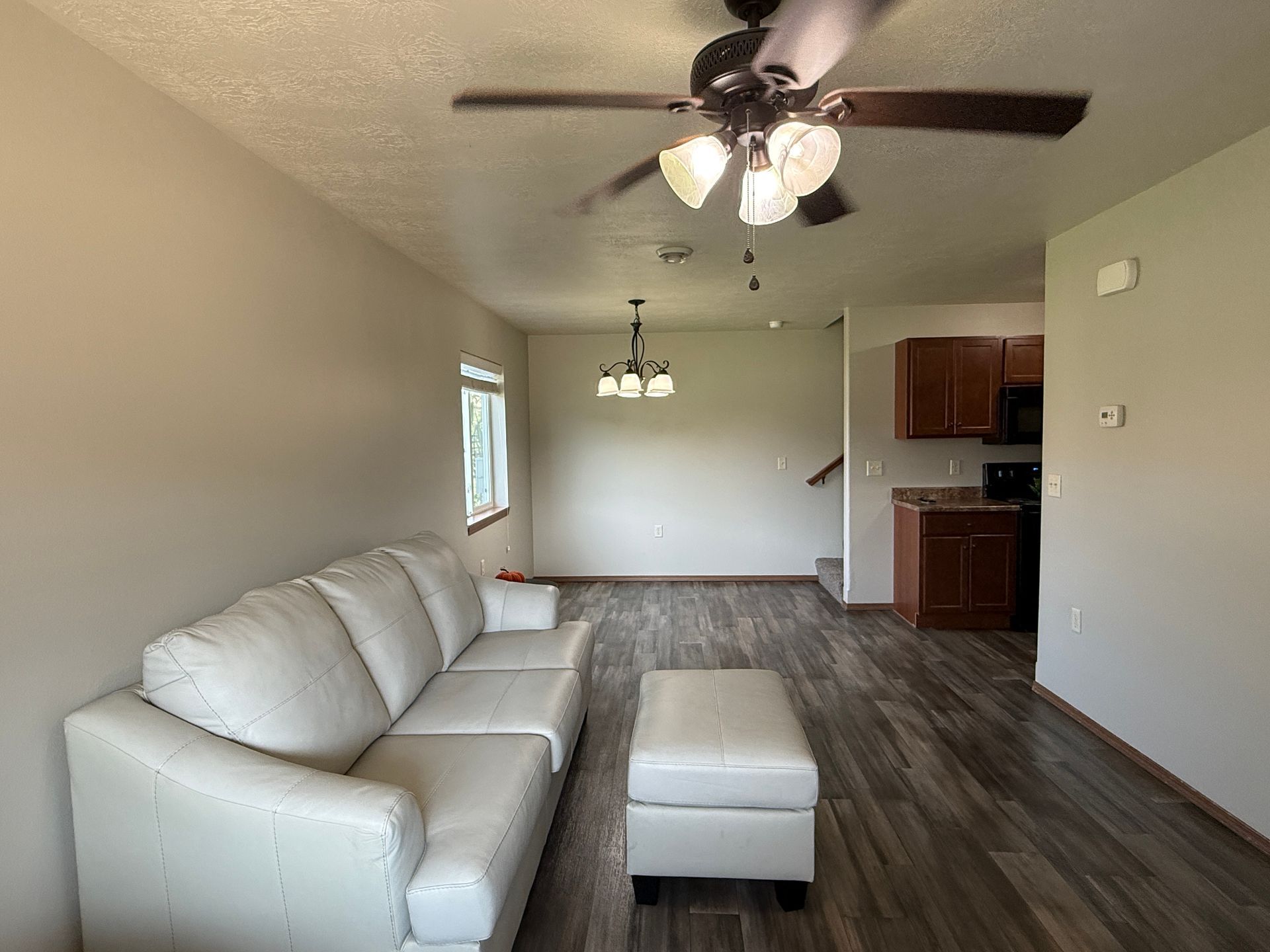 Empty living room with white sofa and ottoman, ceiling fan, wood floors, and a small kitchen area.