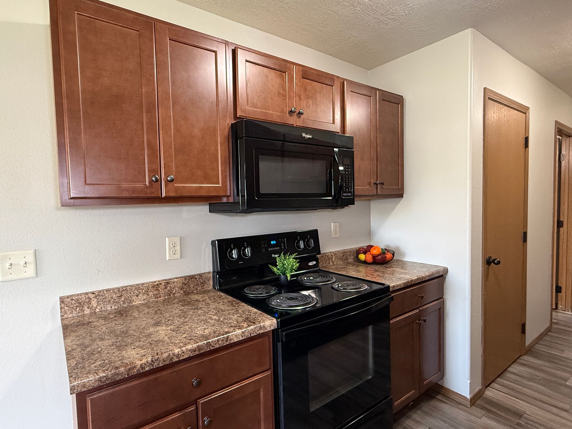 Small kitchen with brown cabinets, black stove and microwave, speckled countertops, and wood-look flooring.