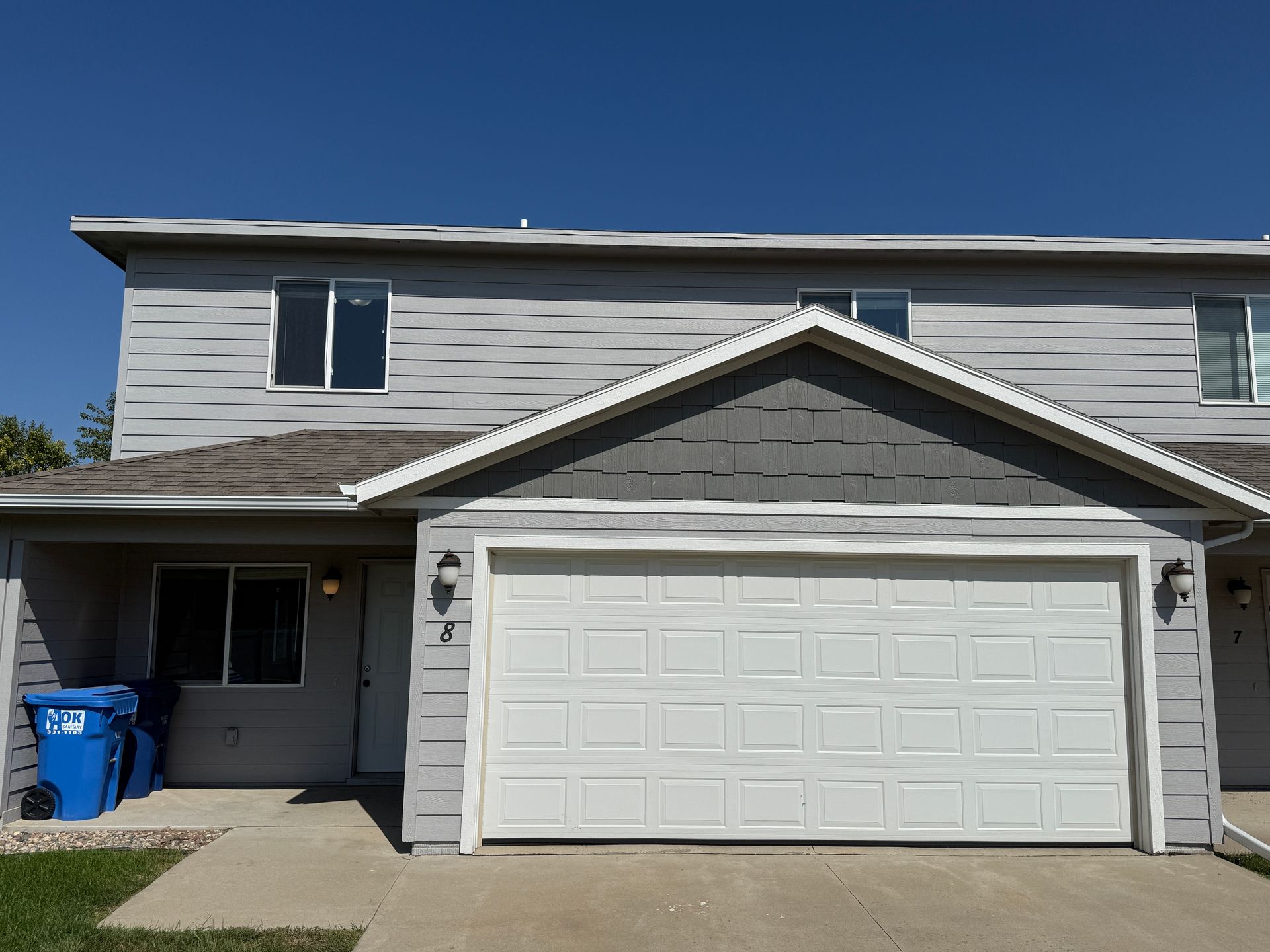 Two-story gray house with a white garage door and a blue recycling bin by the front entrance