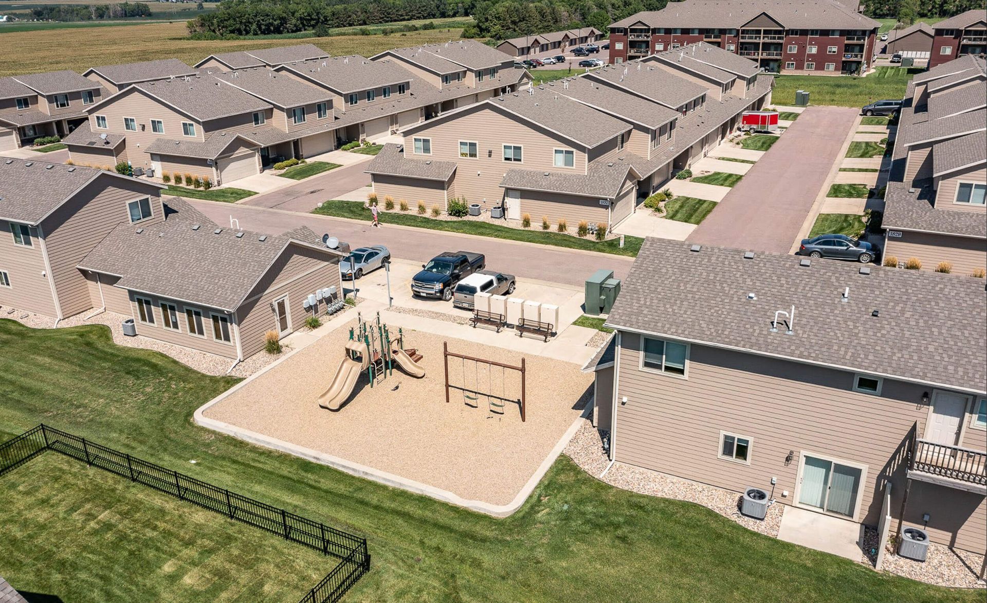 Aerial view of a suburban apartment complex with beige buildings, patios, parked cars, and a playground in the center.