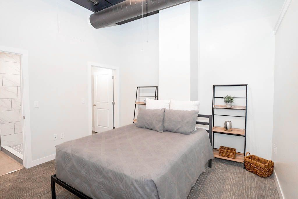 Minimal bedroom with a gray bed, white walls, and a small black shelf next to a wicker basket.