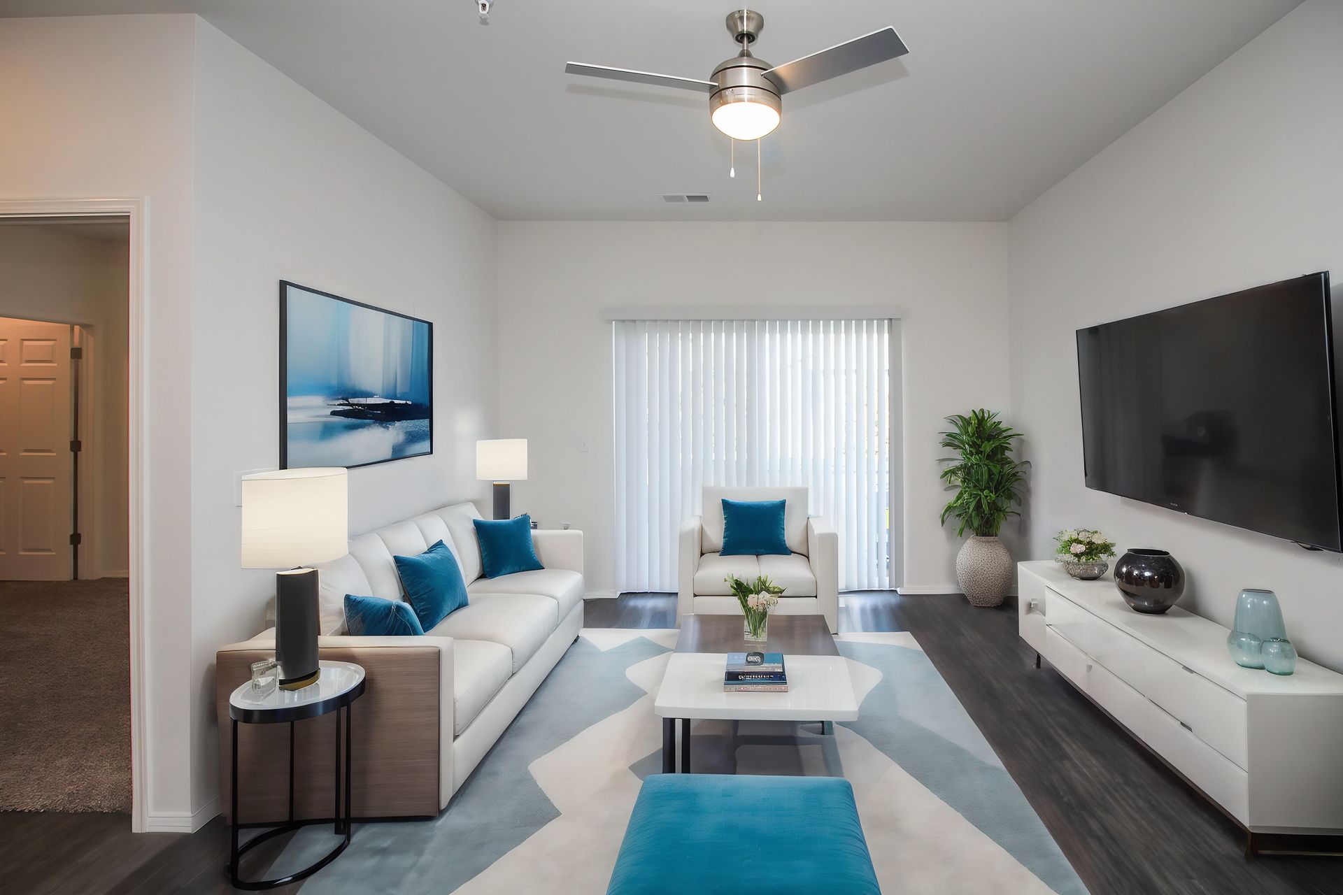 Bright modern living room with white sofa, wall-mounted TV, teal accents, and large window blinds
