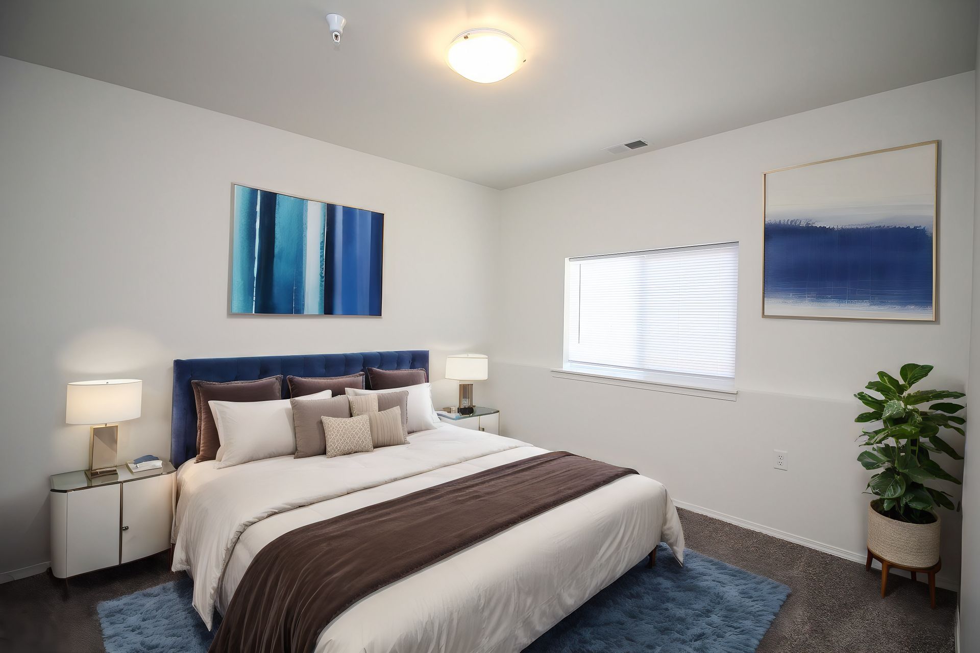 Bright modern bedroom with a made bed, blue artwork, white walls, window blinds, and a potted plant