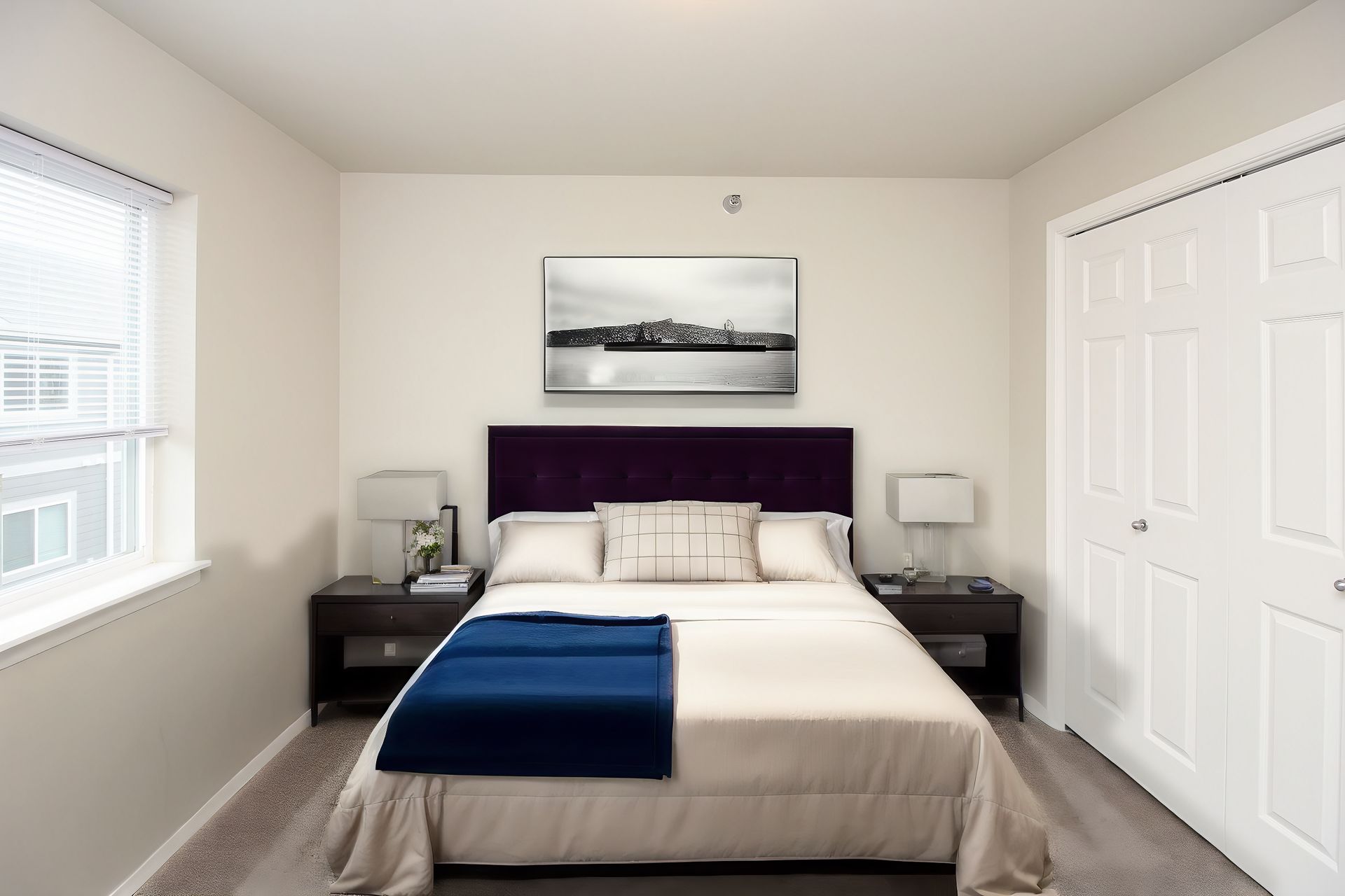 Bright bedroom with a bed, dark nightstands, white closet doors, and a framed wall photo.
