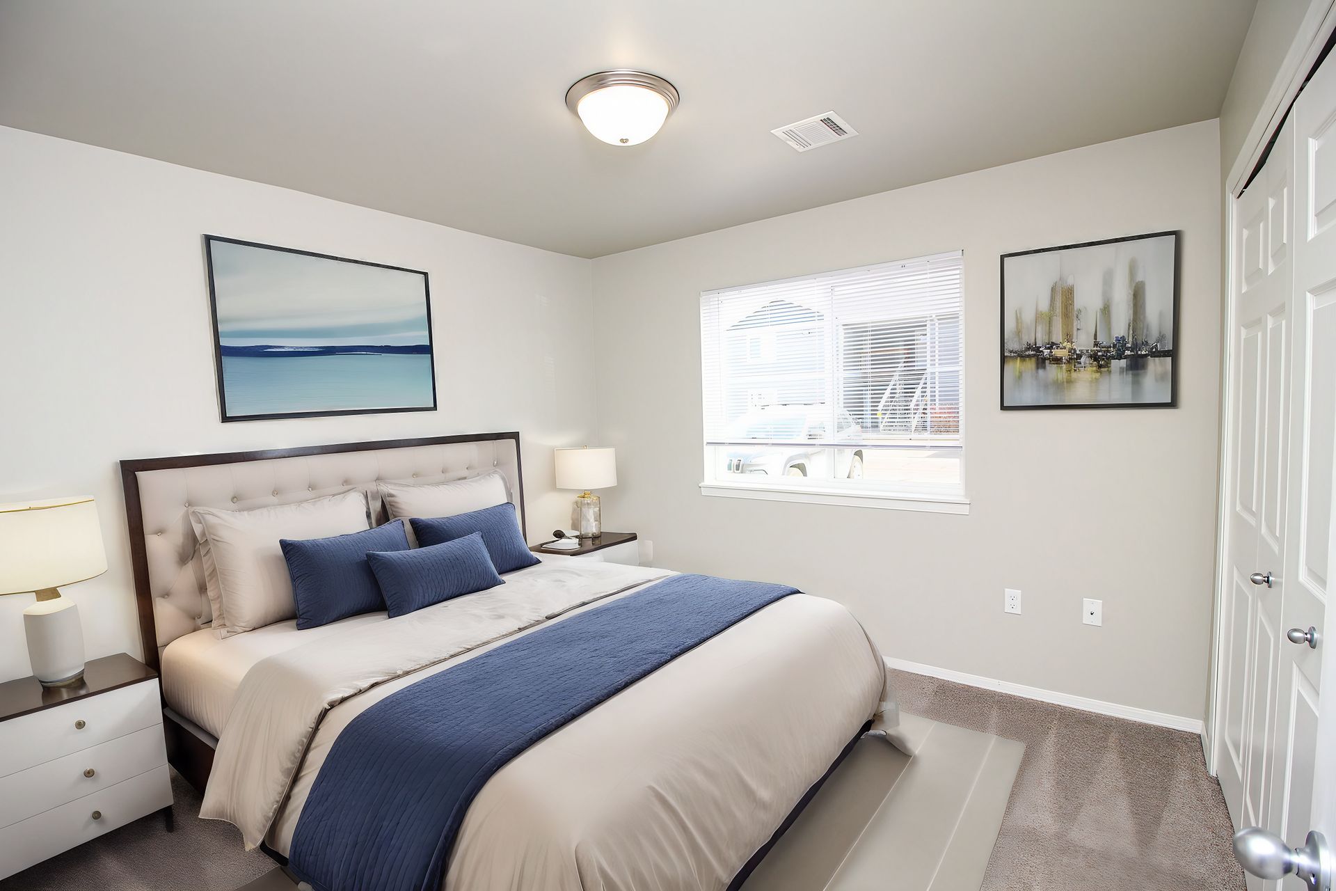 Bright bedroom with a neatly made bed, blue accents, framed art, and a window with blinds.