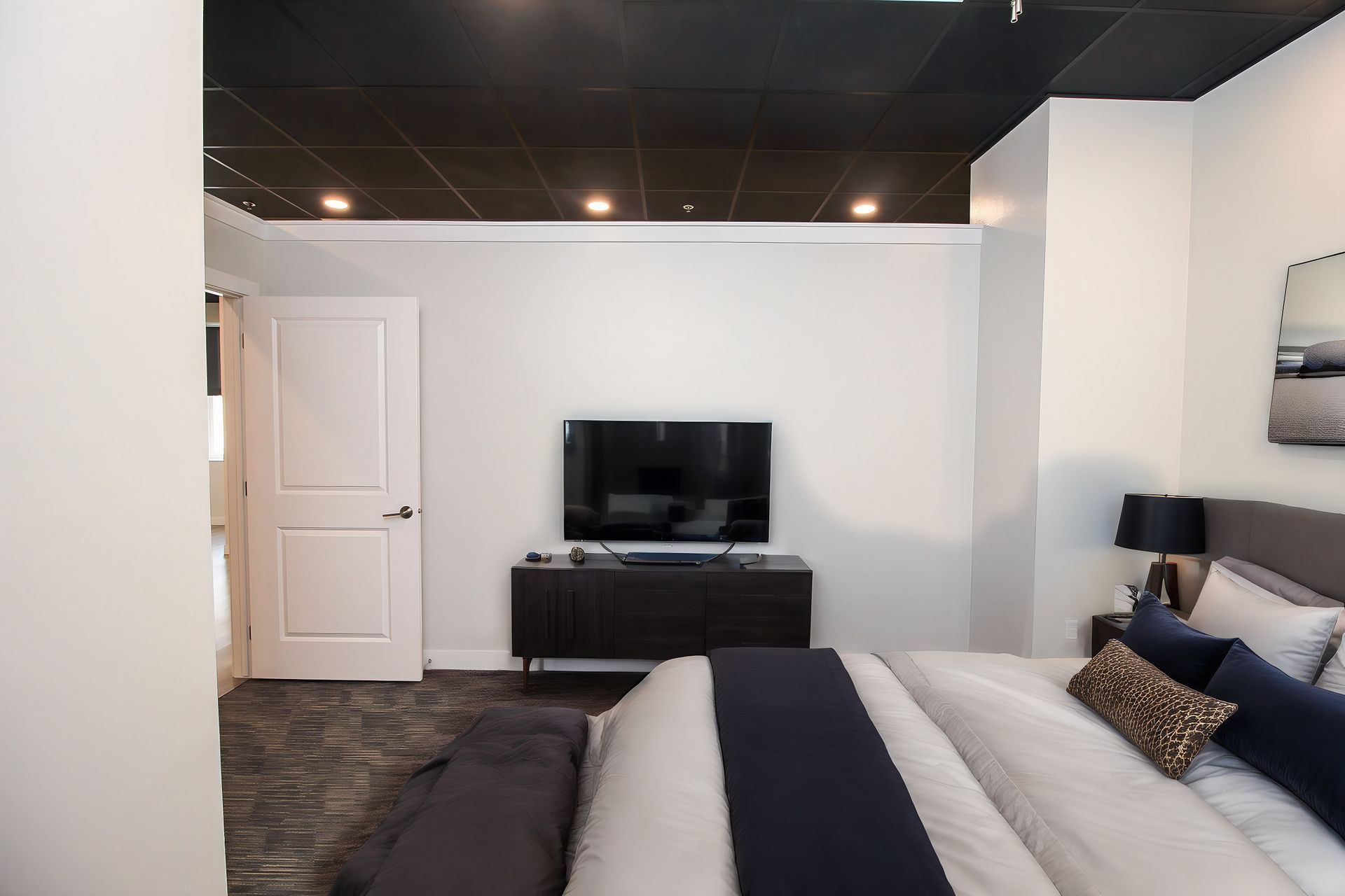 Bedroom with king bed, wall-mounted TV, dresser, white walls, and open door near the ceiling lights.