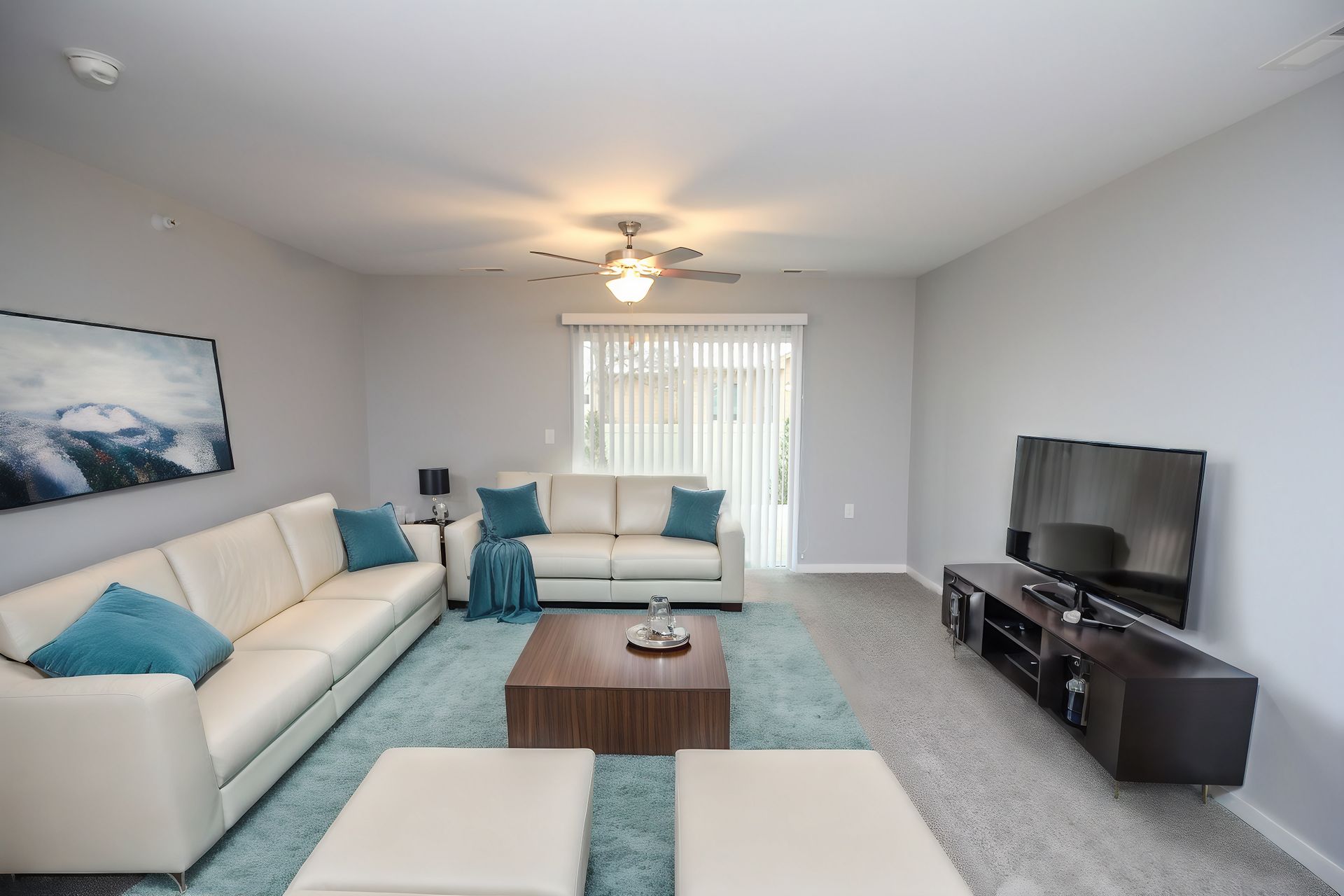 Bright living room with white sofas, teal accents, coffee table, and TV on a stand near a window.
