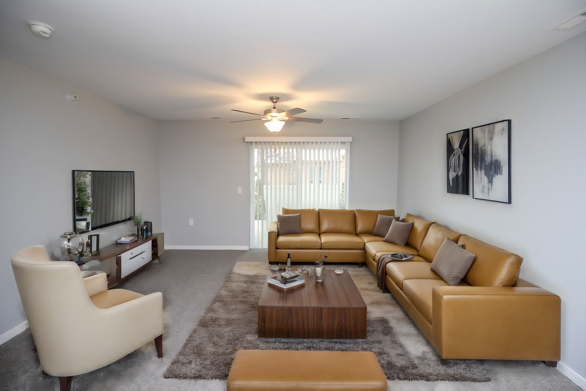 Modern living room with tan sectional, beige chair, coffee table, TV, rug, and sliding door blinds