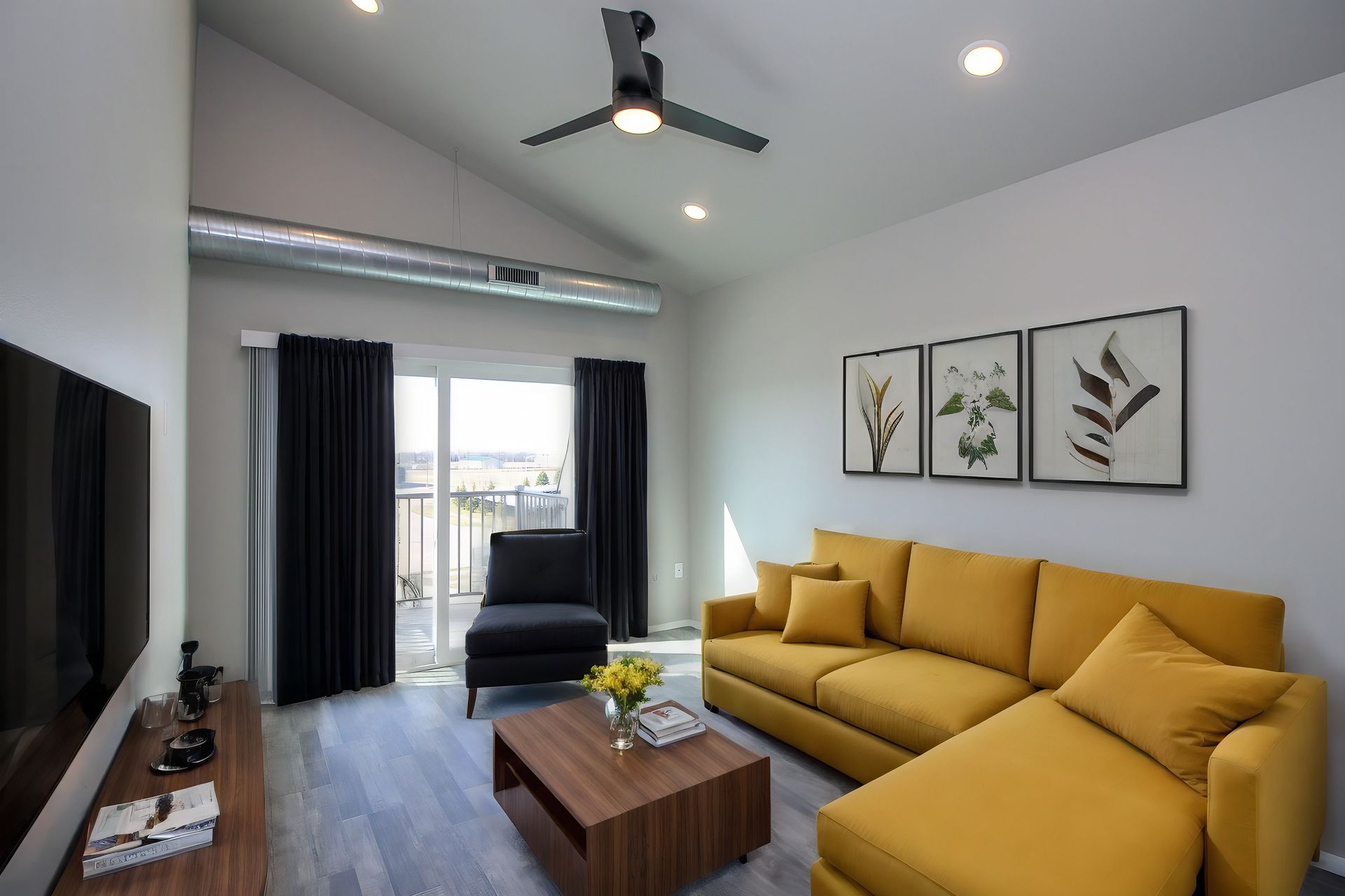 Modern living room with mustard sofa, black chair, coffee table, wall art, and ceiling fan near balcony doors