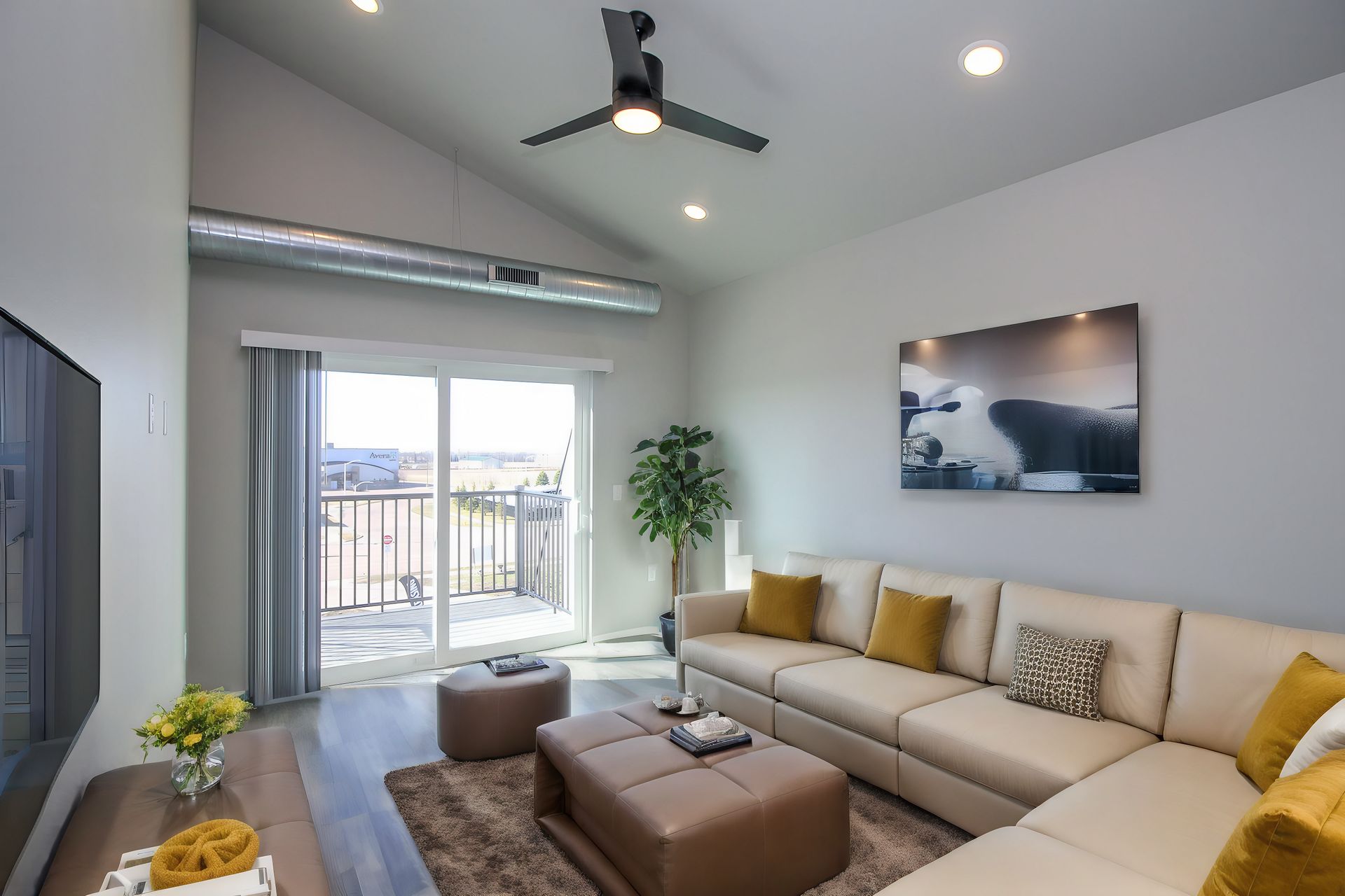 Bright modern living room with beige sectional, ottoman, wall art, balcony doors, and ceiling fan.