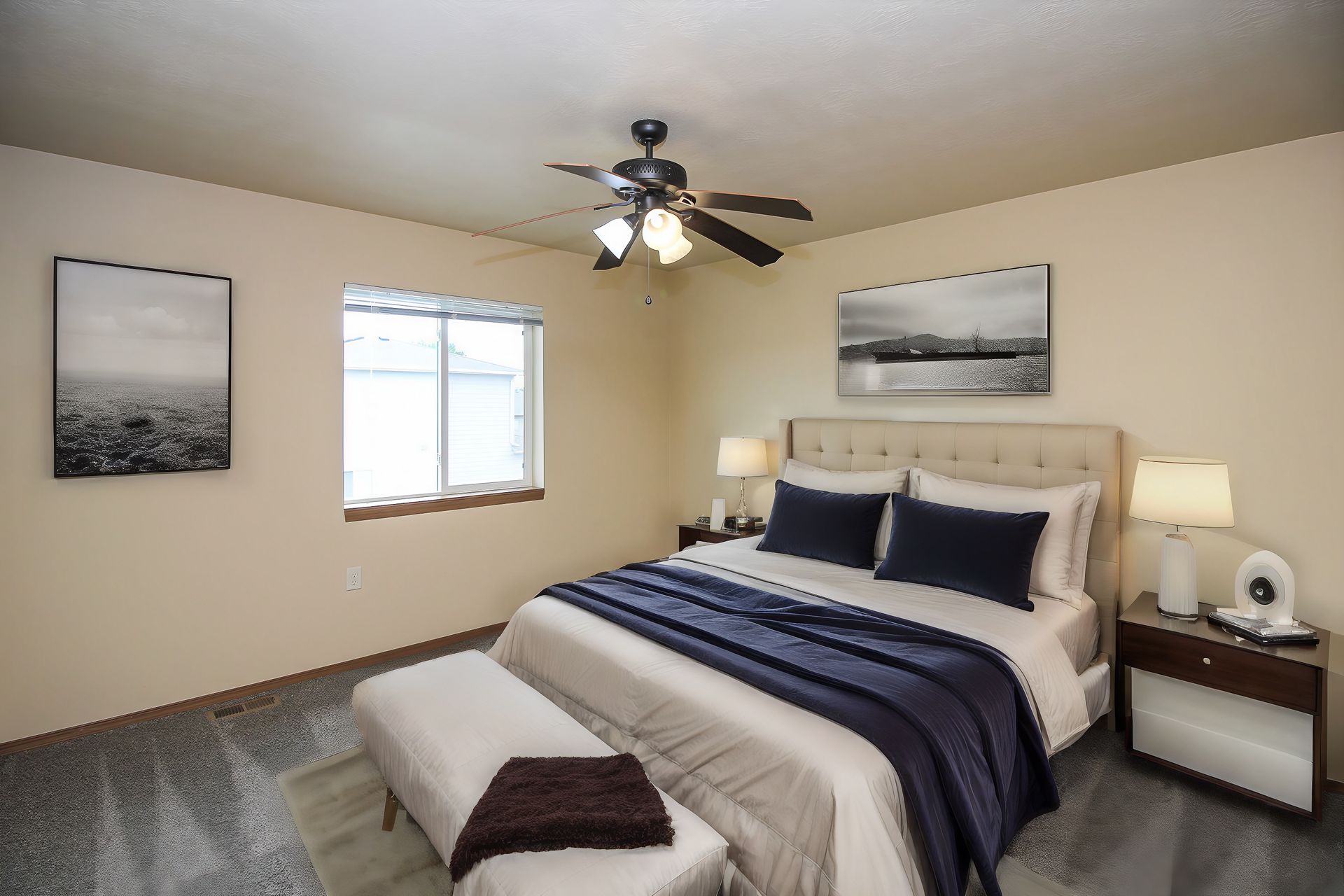 Bedroom with a neatly made bed, beige walls, ceiling fan, and two framed landscape prints.