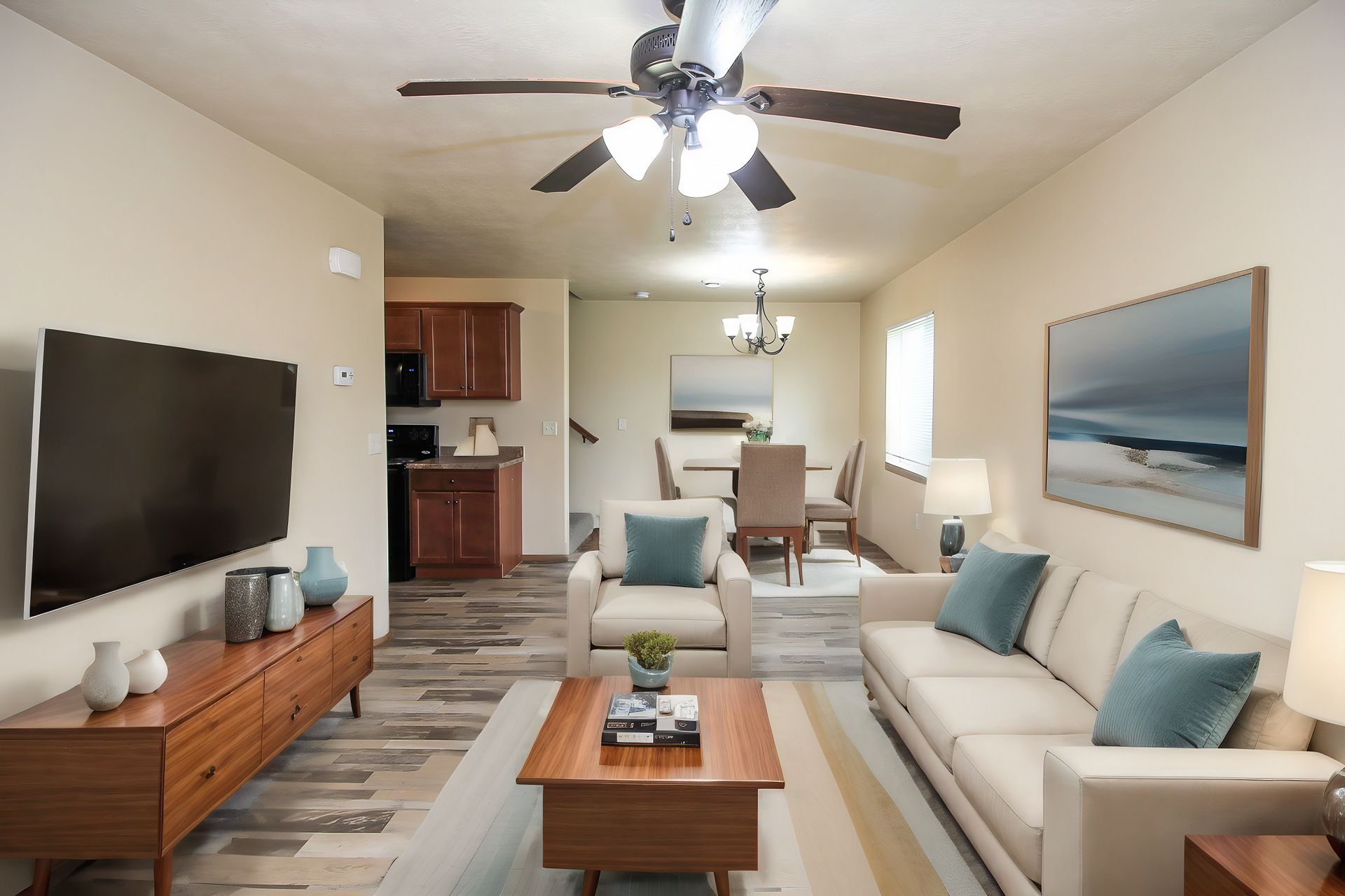 Bright living room with beige sofas, wooden coffee table, wall TV, ceiling fan, and coastal wall art