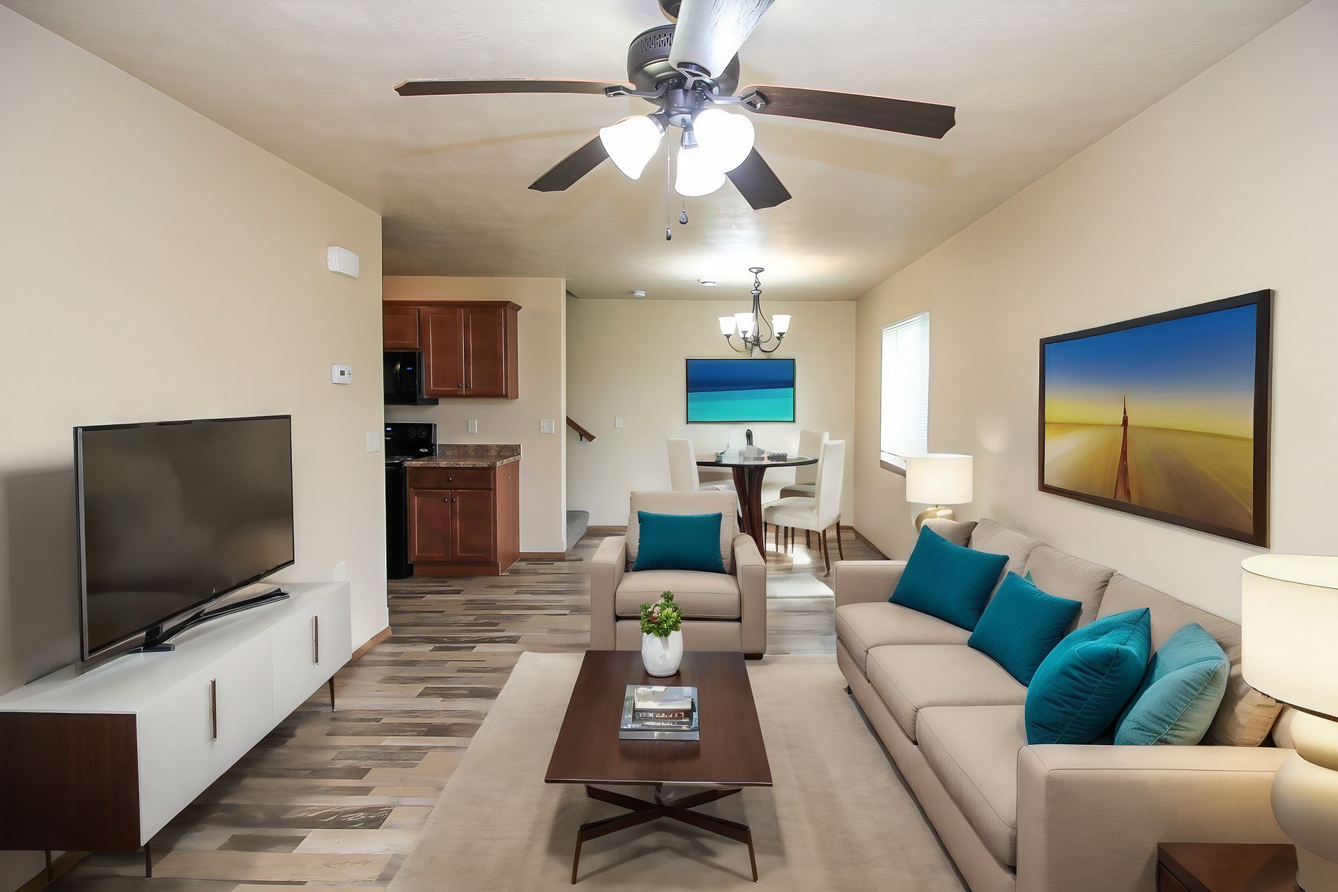 Bright living room with beige sofa, teal pillows, TV, ceiling fan, and wall art under warm lighting
