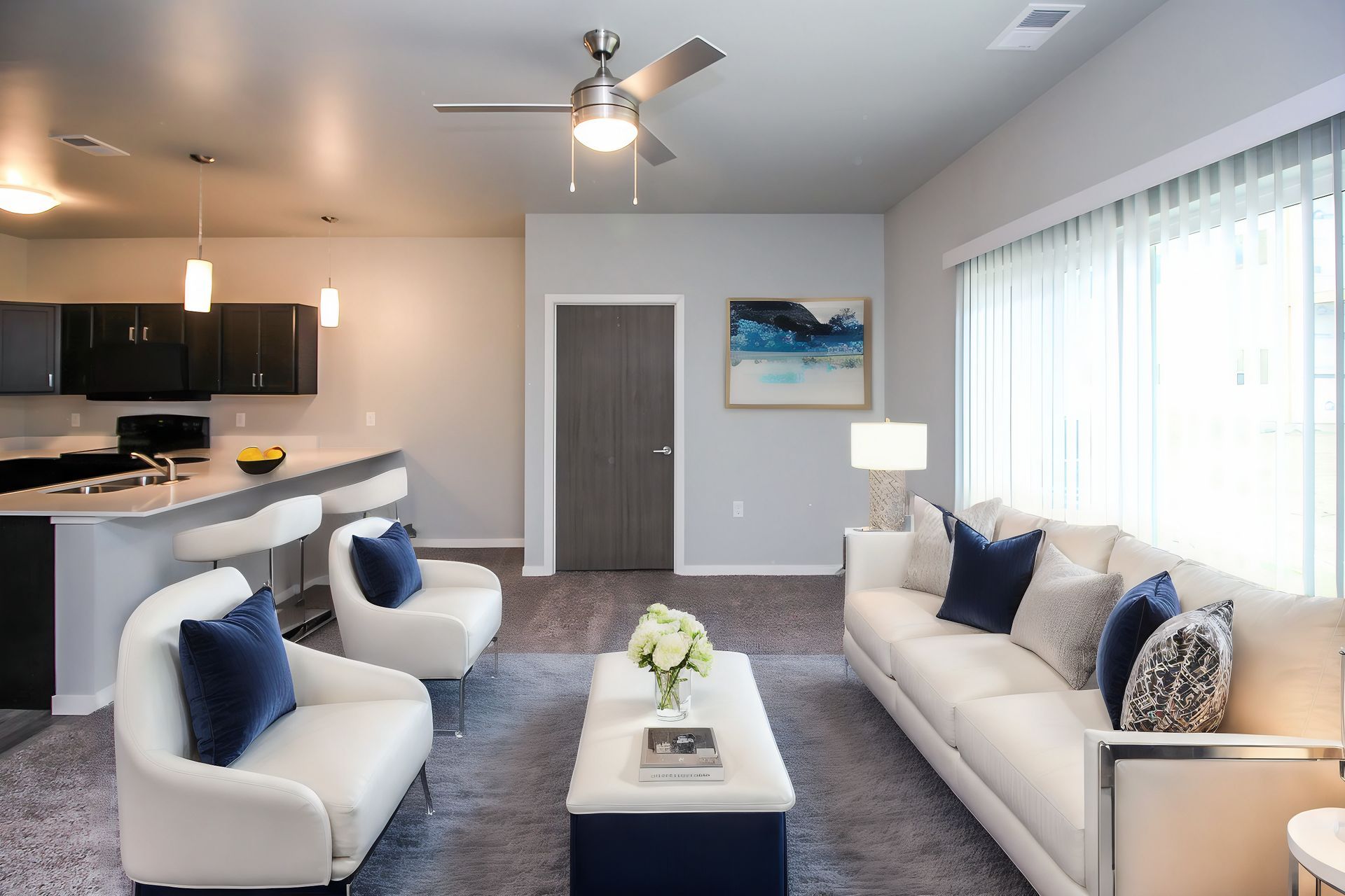 Bright modern living room with white sofas, blue pillows, kitchen bar, and large windows.