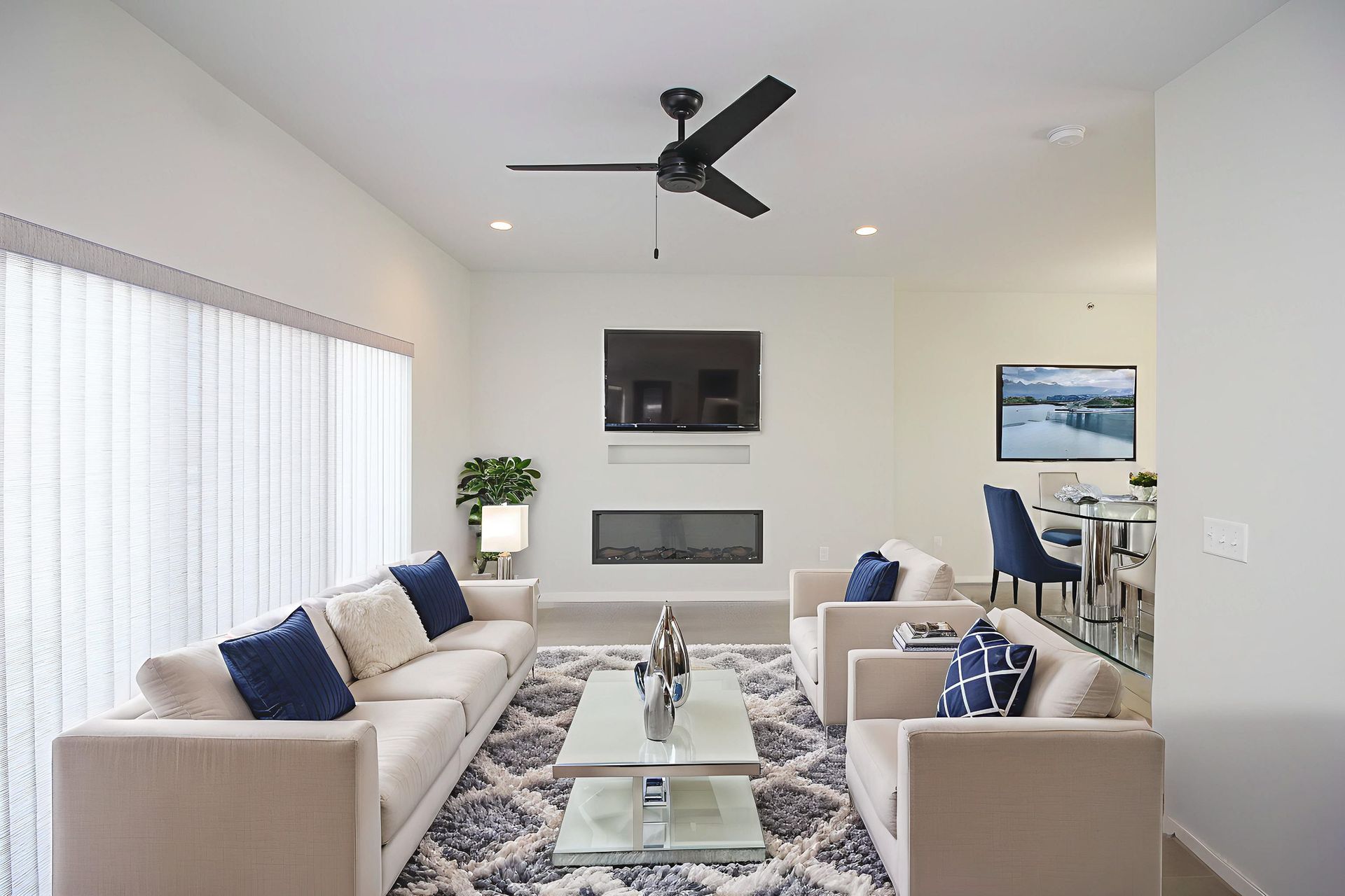 Bright modern living room with white sofas, blue pillows, patterned rug, fireplace, TV, and black ceiling fan