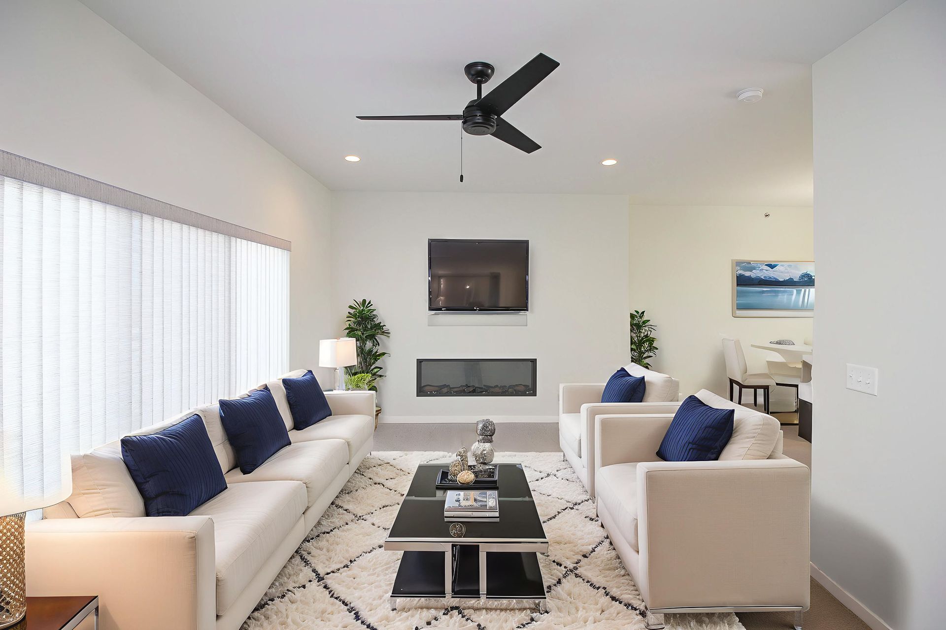 Bright modern living room with white sofas, navy pillows, coffee table, wall TV, and ceiling fan