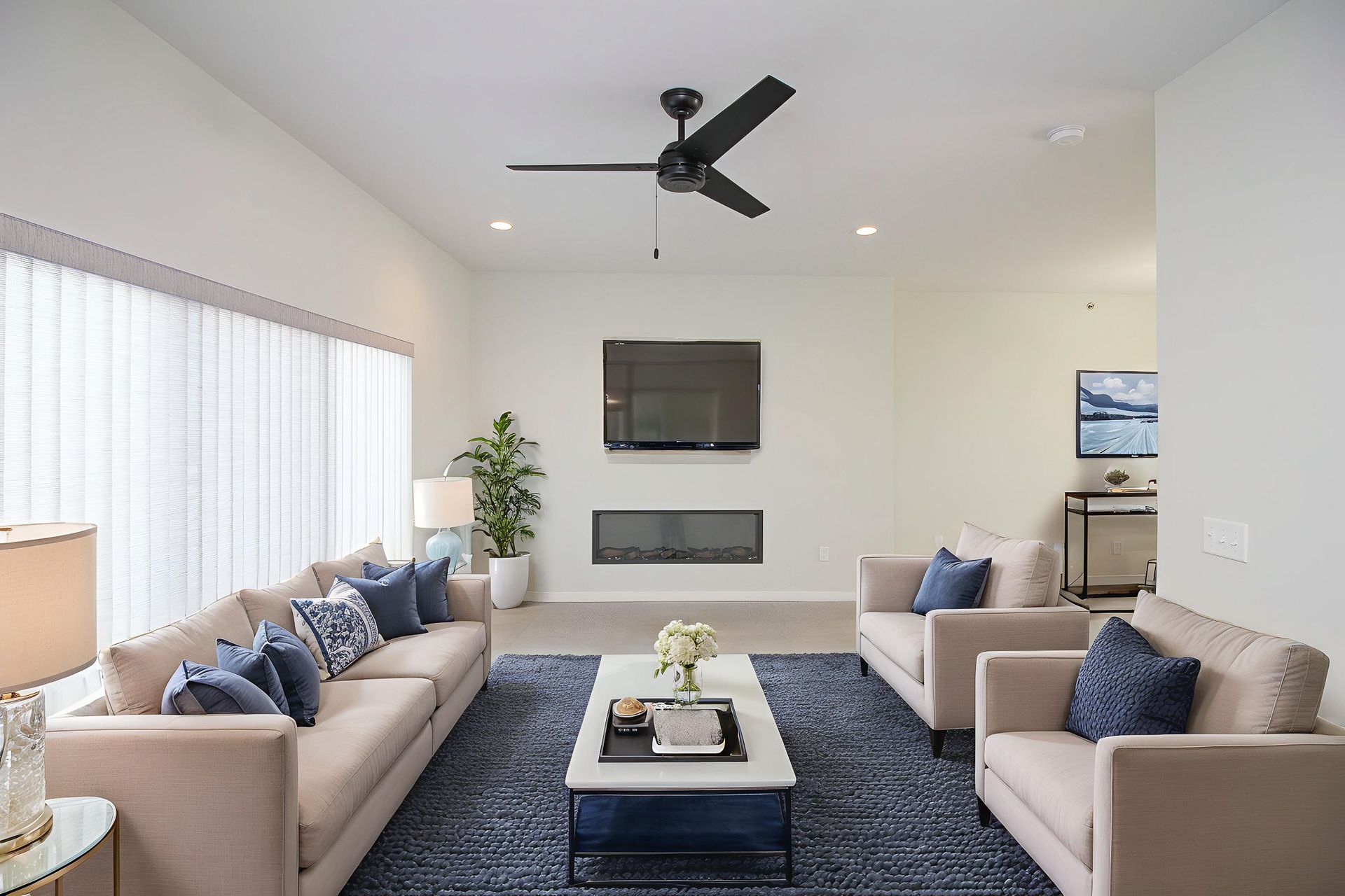 Bright modern living room with beige sofas, blue pillows, wall TV, fireplace, and ceiling fan