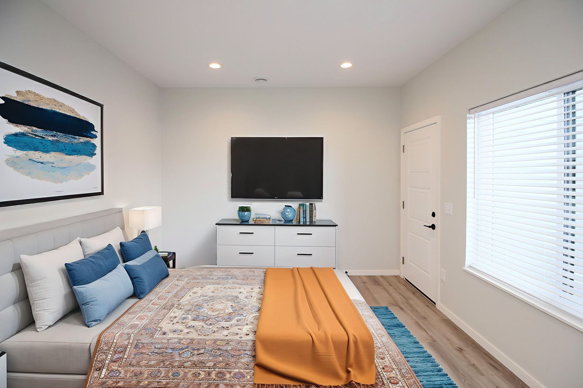 Modern bedroom with a bed, dresser, wall-mounted TV, blue accents, and bright window blinds.