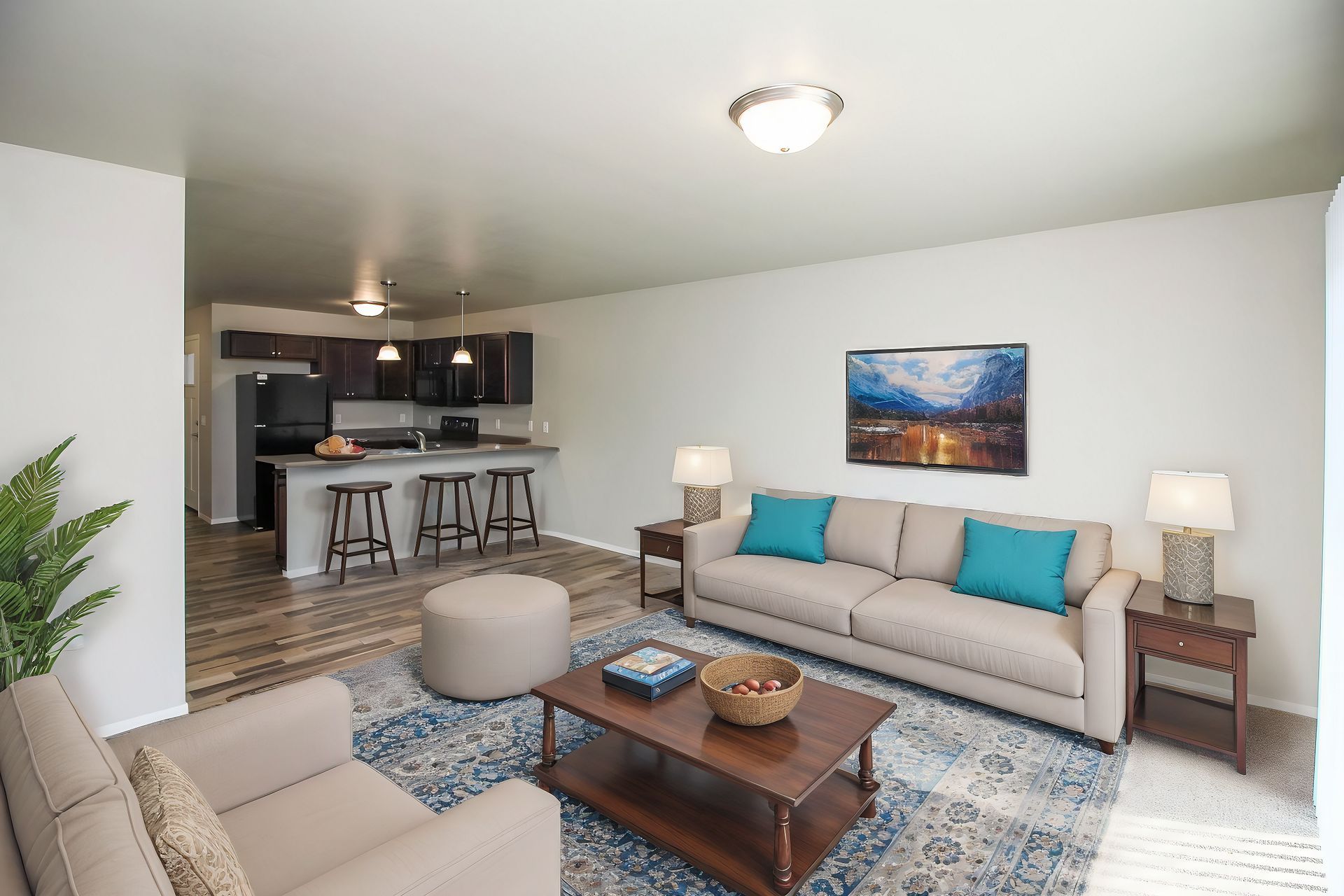Bright living room with beige sofa, blue accents, coffee table, and open kitchen in the background