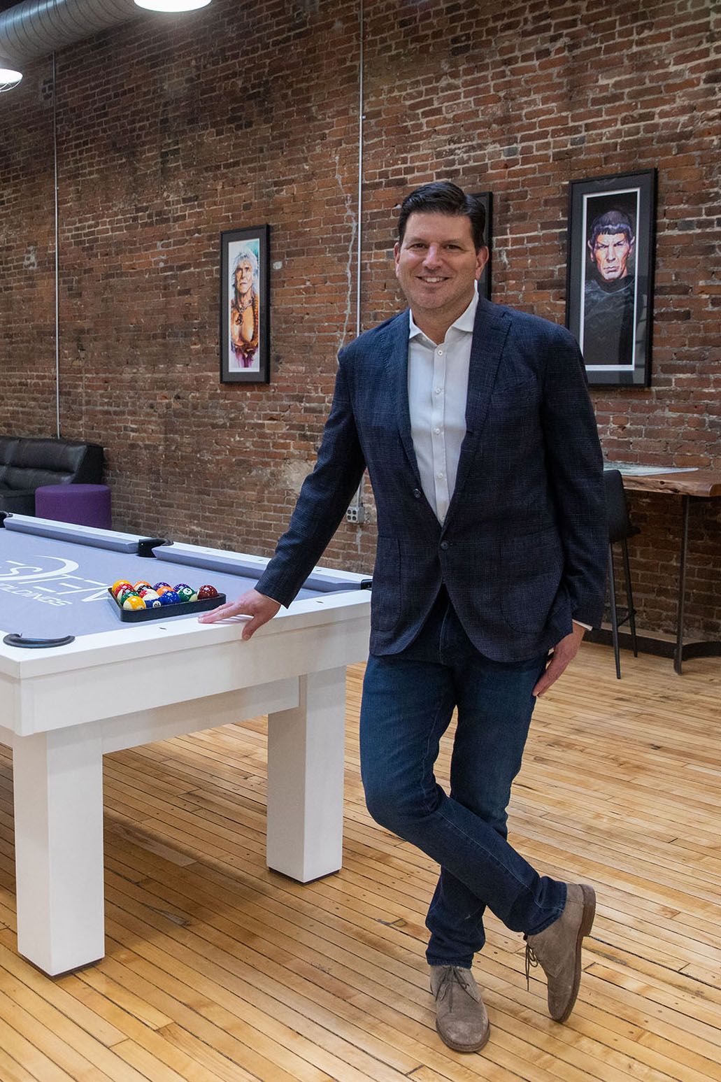 Man in blazer leaning on a white pool table in a brick-walled room with framed posters.