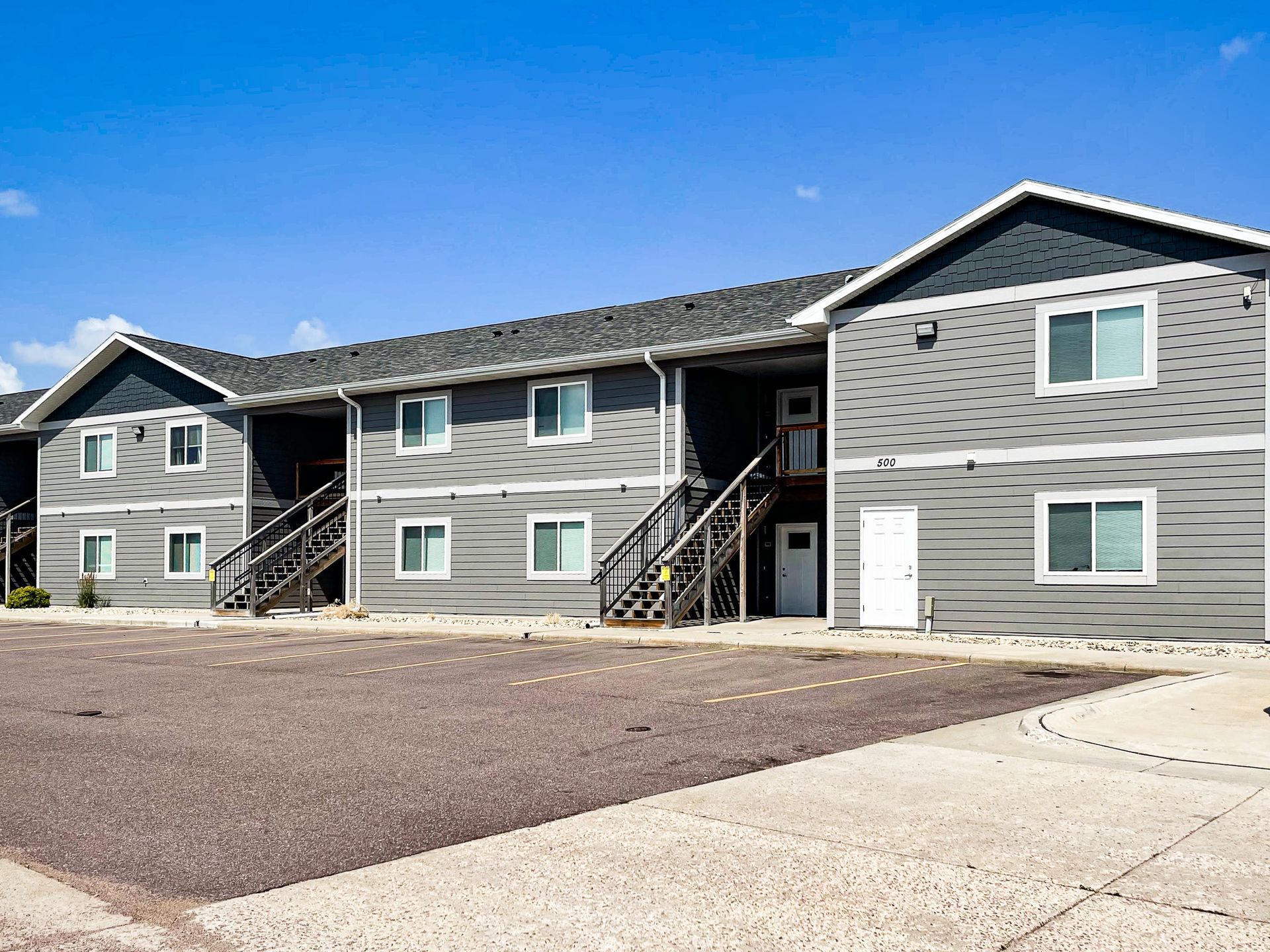 Gray apartment buildings with exterior stairs and white trim on a gravel lot under a blue sky