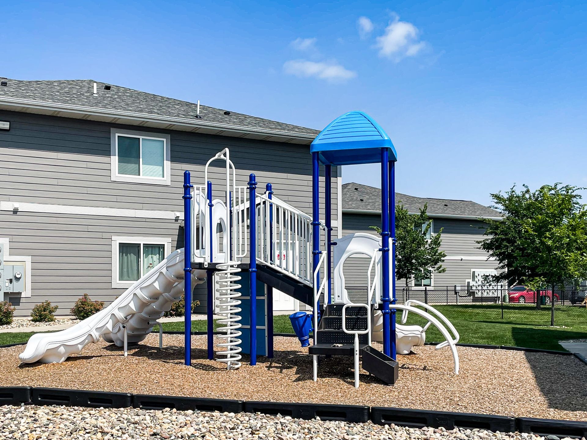 Blue playground structure with slides and climbing bars in front of apartment buildings under a clear sky