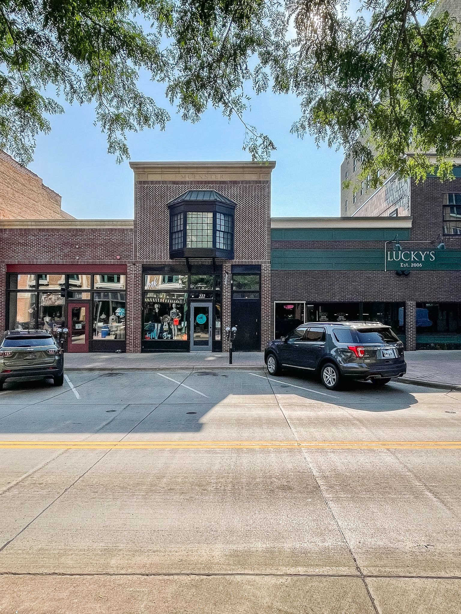 Storefronts in a shopping plaza with parked cars and trees overhead