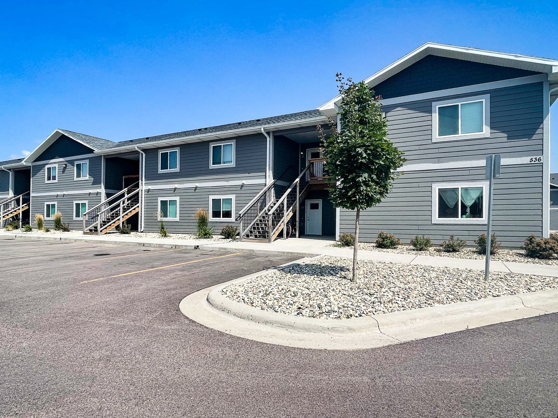 Gray apartment building exterior with blue trim and a gravel parking lot in front
