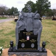 A grey granite monument featuring a statue of the Holy Family over a black plaque with names and photos in a cemetery.