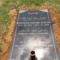 A black granite gravestone set in dirt, inscribed with the name Suleiman Khalid Kullab and his dates of birth and death.