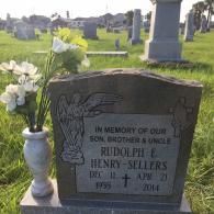 A granite headstone for Rudolph E. Henry-Sellers, dated 1955-2014, with a vase of artificial flowers in a cemetery.
