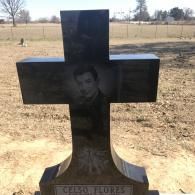 A dark stone cross gravestone for Celso Flores, featuring an etched portrait, situated in an open, grassy field.