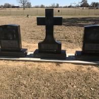 Three black granite headstones, including one in the shape of a cross, sit on a concrete base in a grassy field.