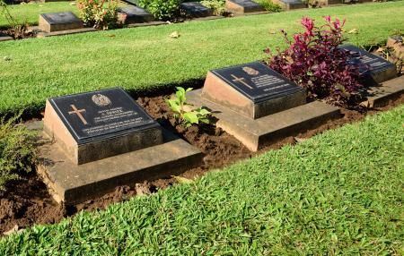 Two stone grave markers with crosses and inscriptions set in a green, grassy cemetery lawn with a small purple bush.