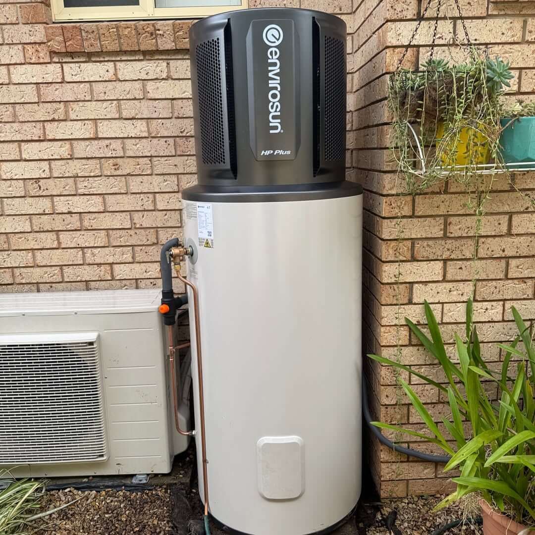 A white Envirosun HP Plus water heater is sitting next to a brick wall.