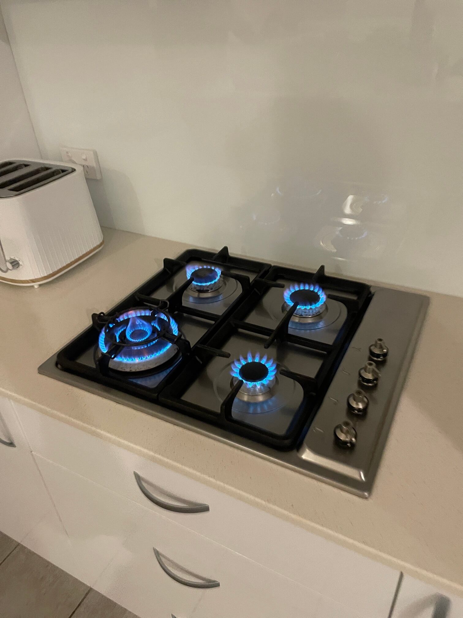 A stainless steel gas stovetop with four burners ignited, showing blue flames, set on a kitchen counter.