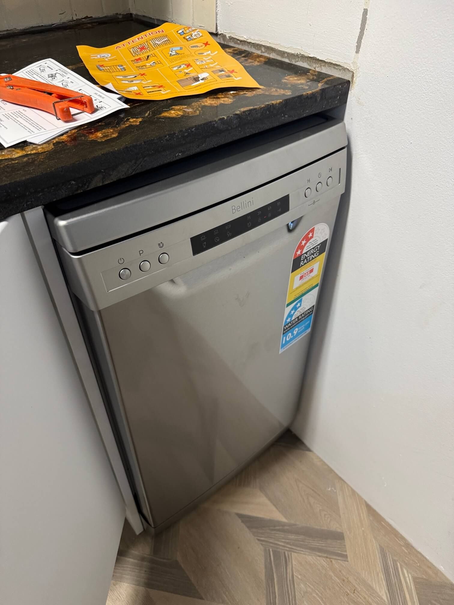 A stainless steel dishwasher installed under a dark countertop in a kitchen with herringbone-patterned flooring.