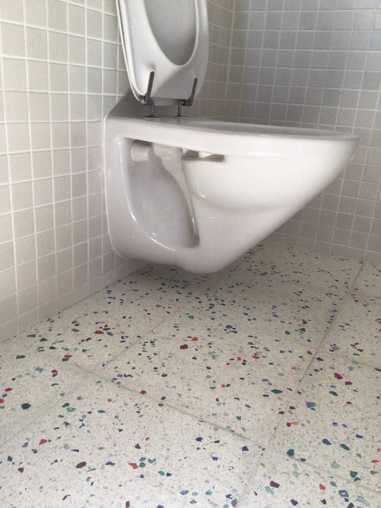 A white wall-mounted toilet in a bathroom with white tiled walls and a multi-colored speckled tile floor.