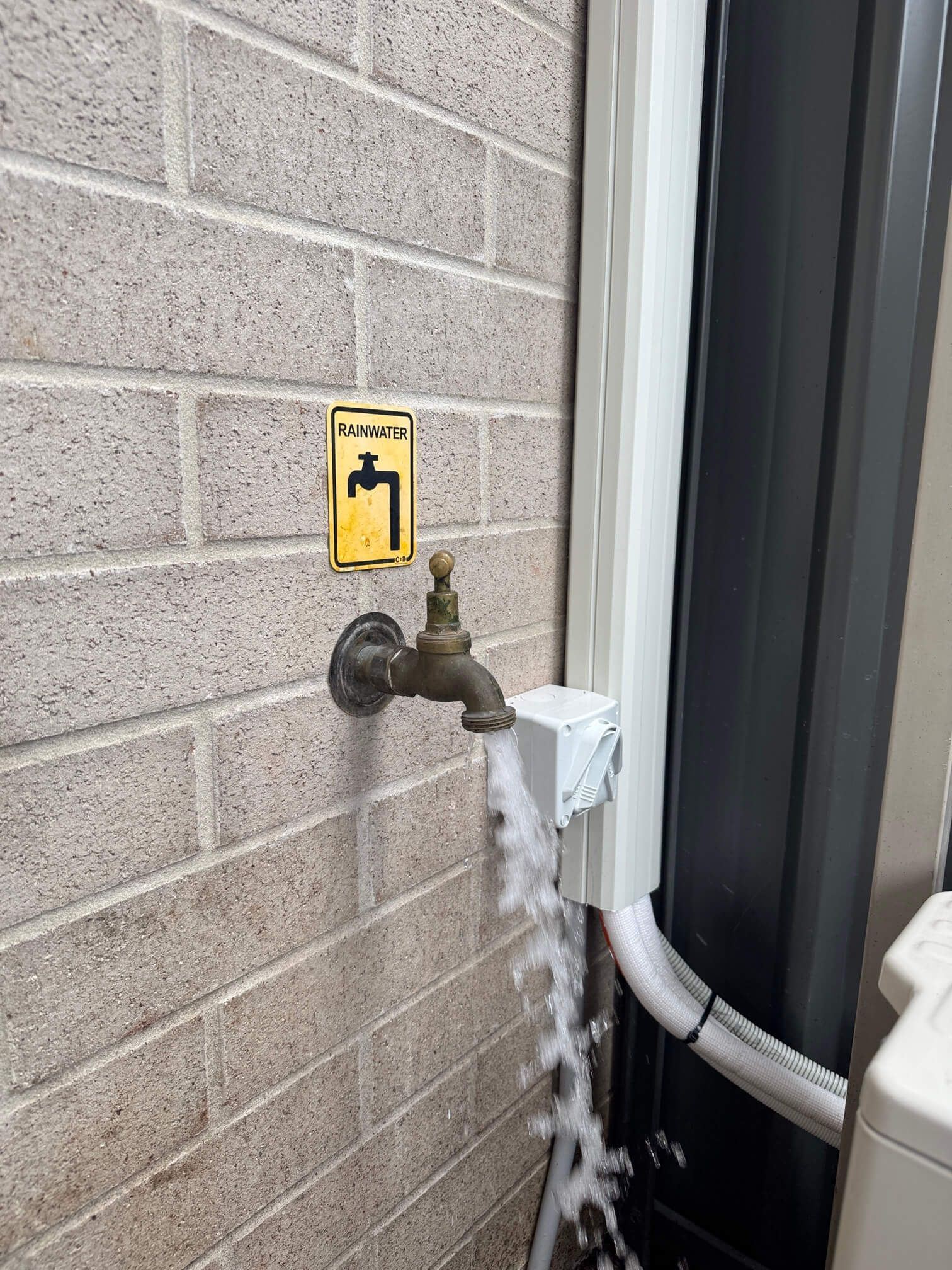 A brass outdoor faucet mounted on a brick wall is actively running water, flowing down directly over an electrical outlet.